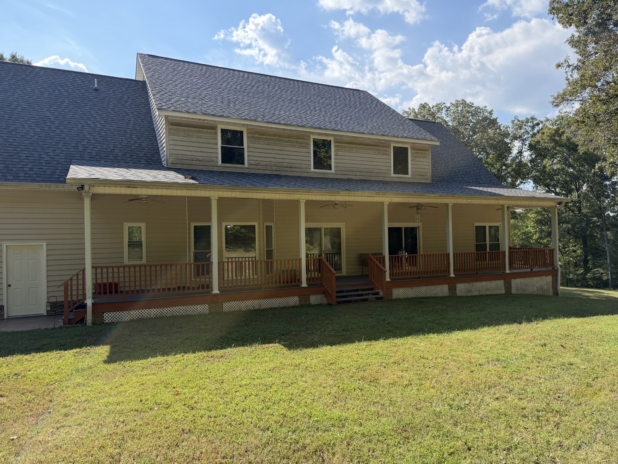 Two-story beige house with a long covered porch and wooden railings overlooking a sunny lawn.