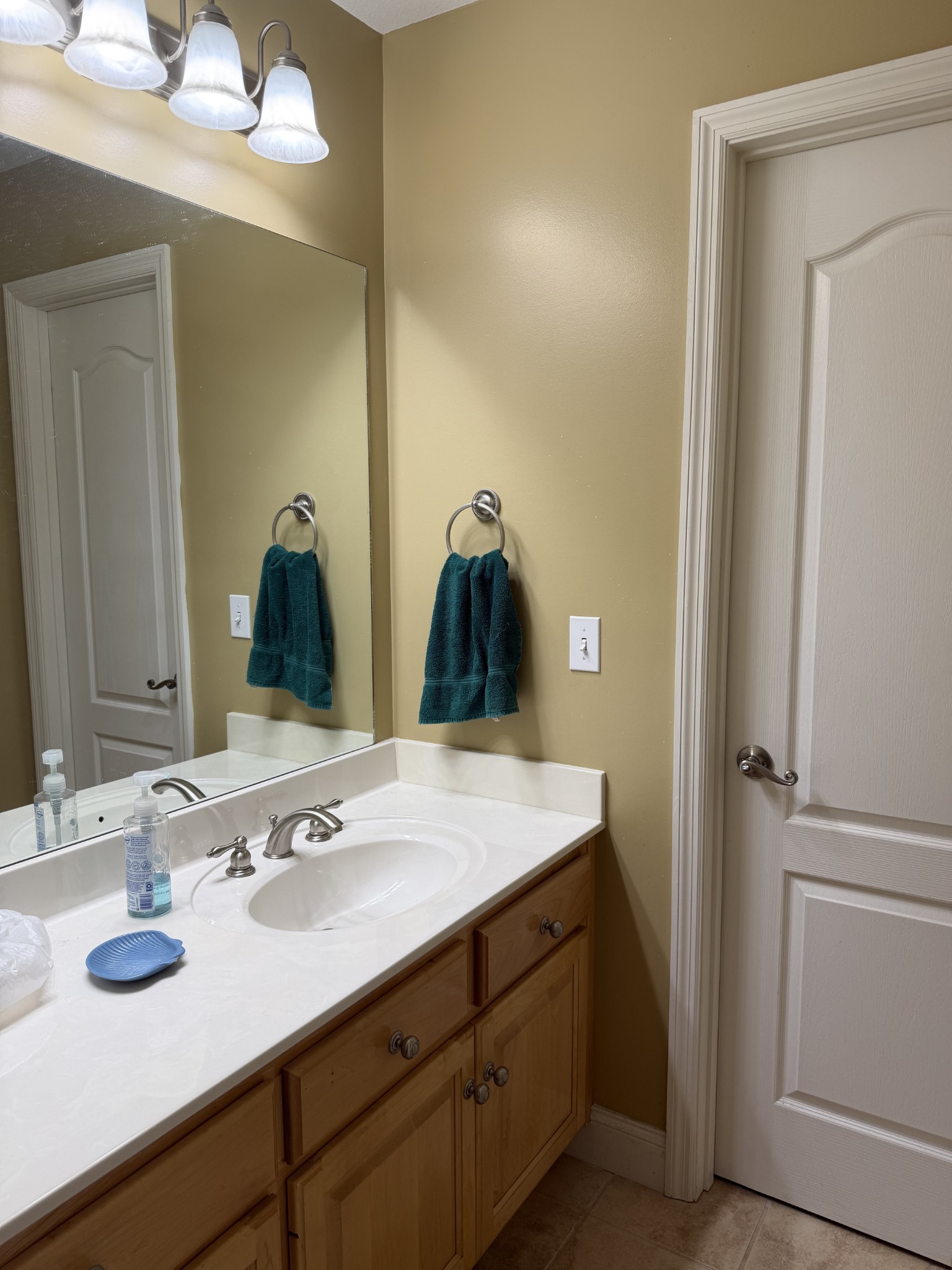 Bathroom vanity with an oval sink, wood cabinets, large mirror, and teal hand towels.