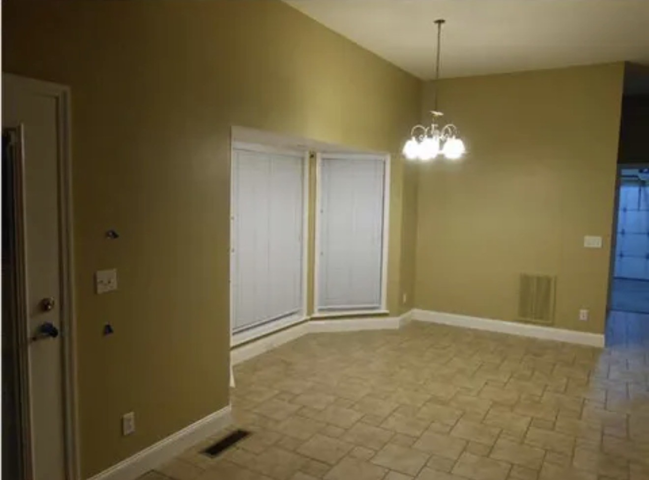 Empty dining area with patterned tile flooring, chandelier, and corner windows with white blinds.