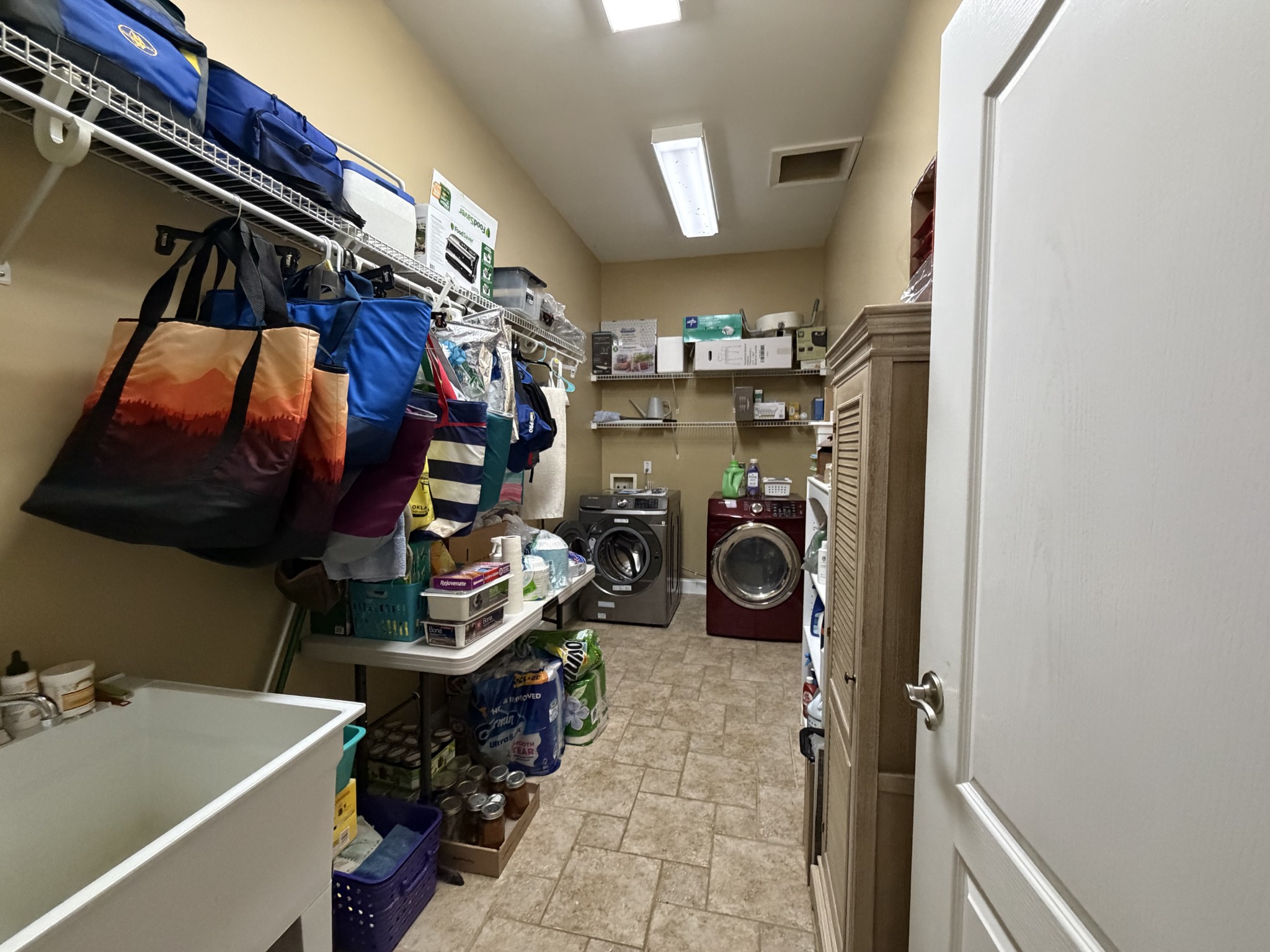 Laundry room with front-loading washer and dryer, crowded storage shelves, and tiled floor.