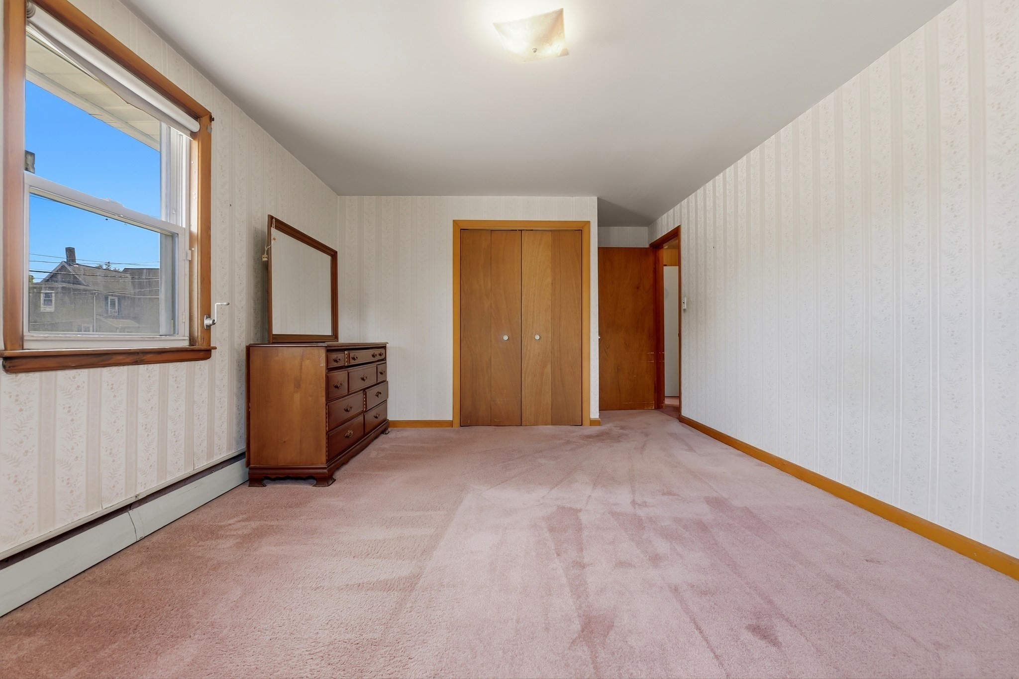 Bedroom with pink carpet, a wooden dresser, and a double-door closet beside a large window.