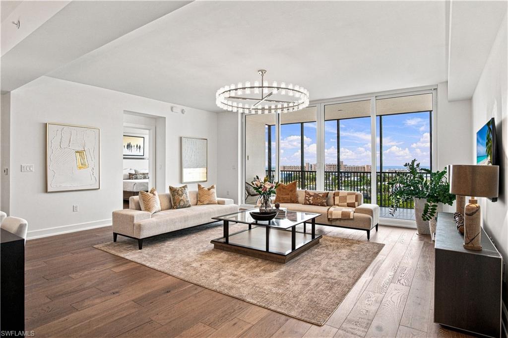 Furnished living room with a glass coffee table, circular chandelier, and balcony overlooking the water.