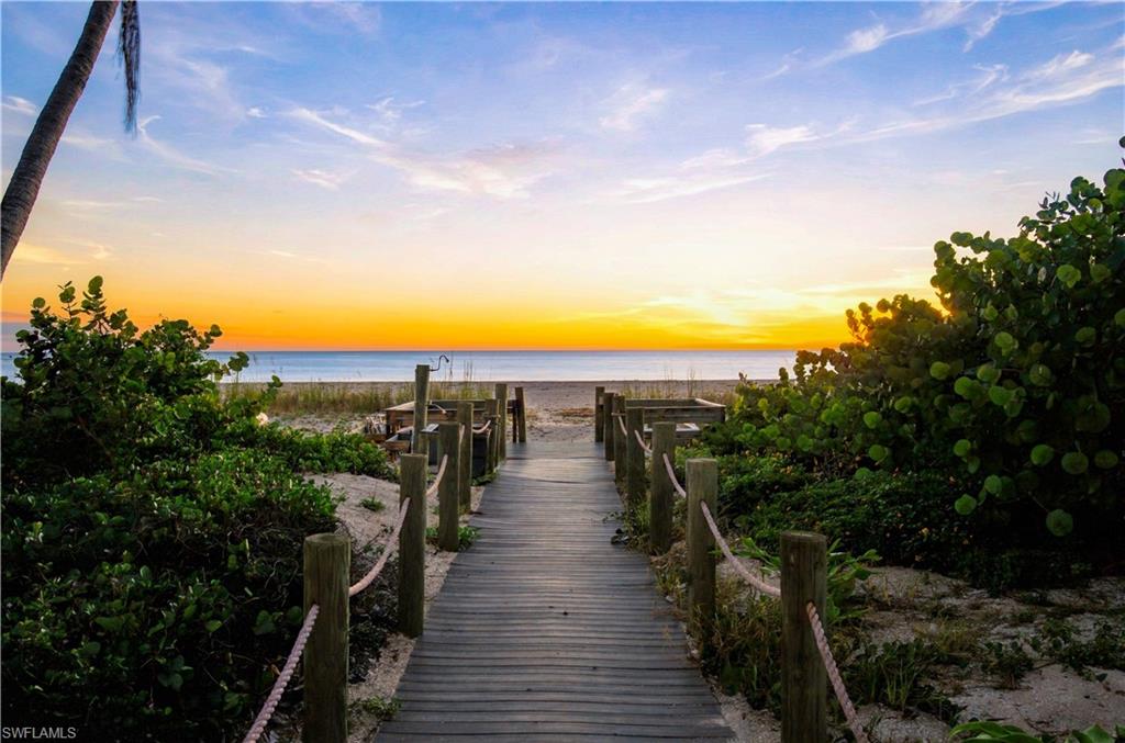 Beach boardwalk framed by coastal greenery leads to the water beneath a glowing sunset.