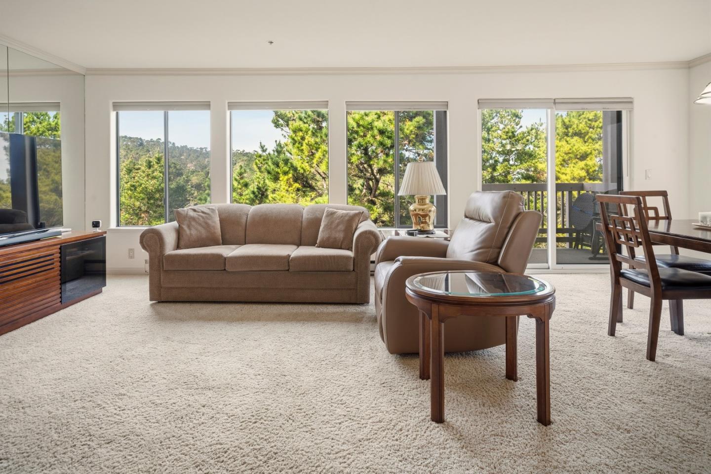 Furnished living room with a beige sofa, leather recliner, and large windows overlooking trees.