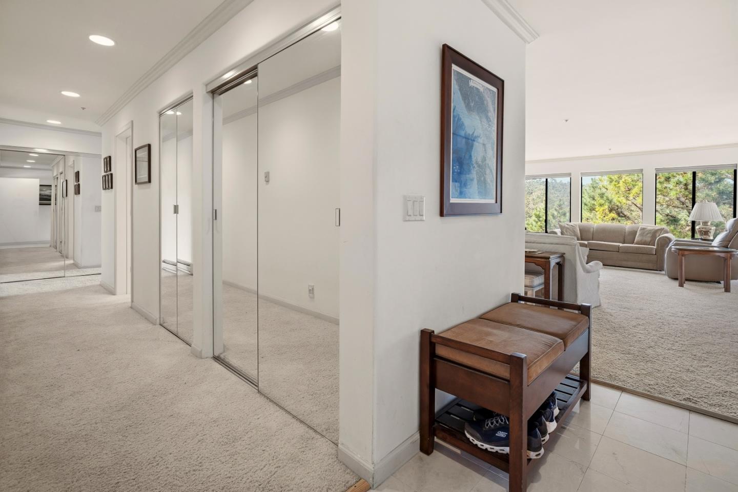 Carpeted entry hall with mirrored closet doors and a wooden shoe bench, opening to a bright living room.