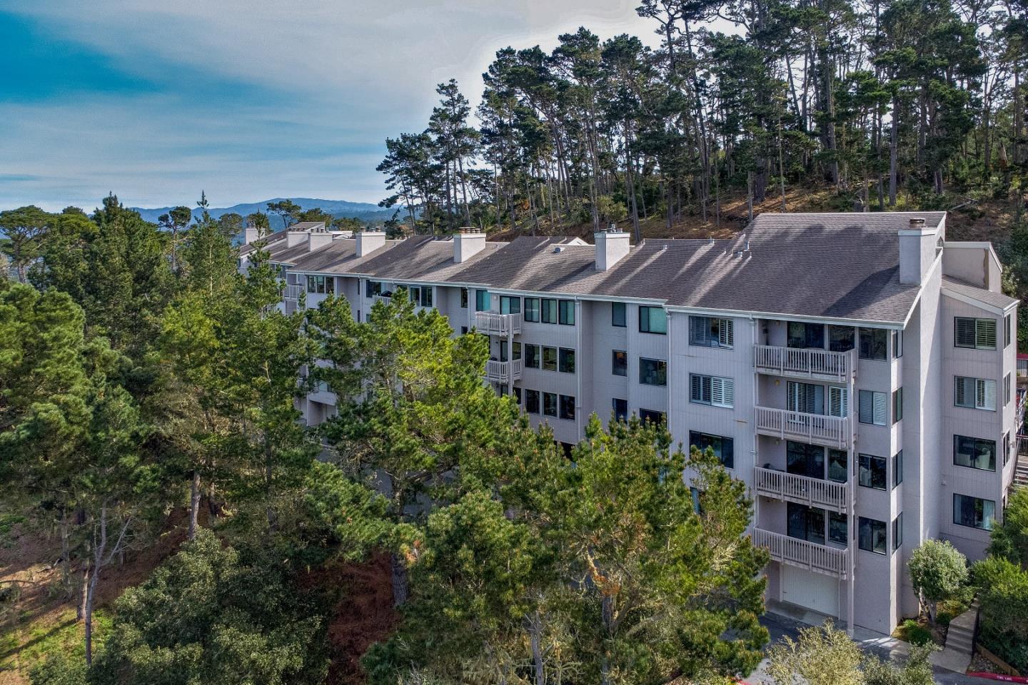 Aerial view of a multi-story condominium building among pine trees, with hills in the distance.