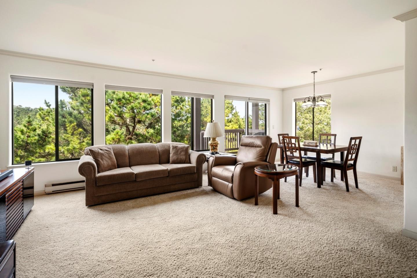 Living and dining room with brown seating and large windows overlooking pine trees.