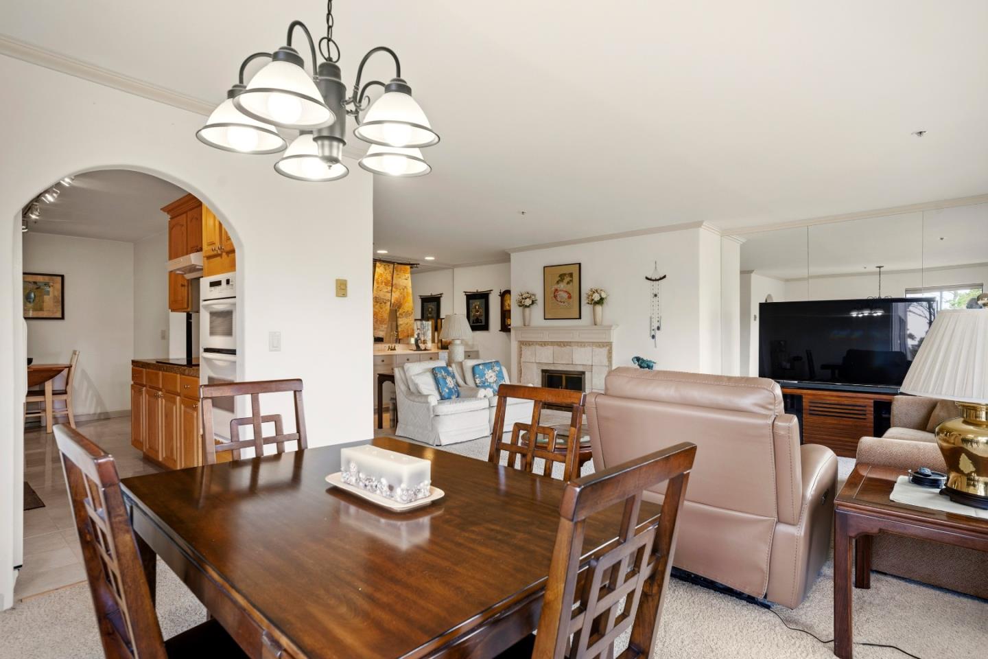 Open-plan dining and living room with a dark wood table, arched kitchen opening, and stone fireplace.