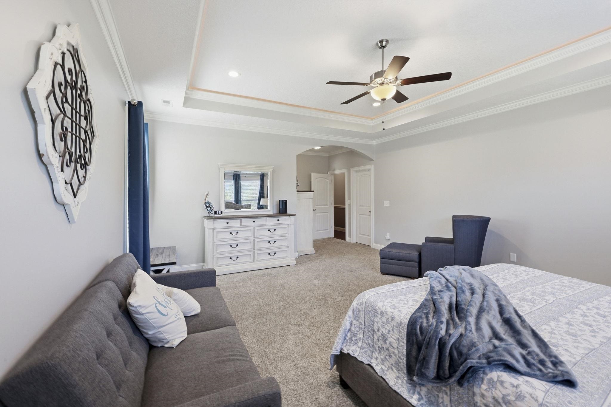 Bedroom with a tray ceiling, ceiling fan, bed, gray sofa, and blue accent chair.