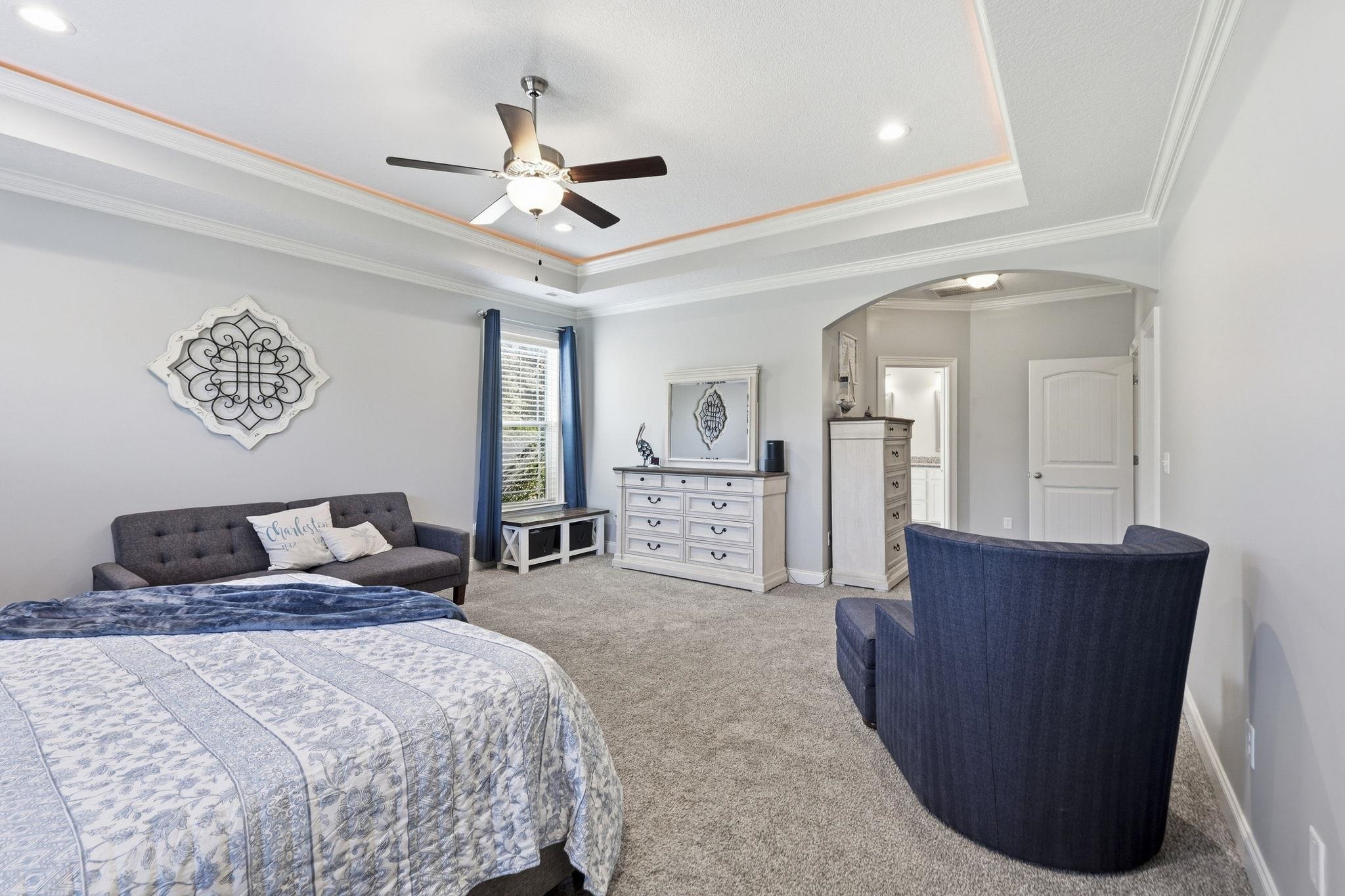 Bedroom with a tray ceiling, ceiling fan, patterned bedspread, and blue seating.