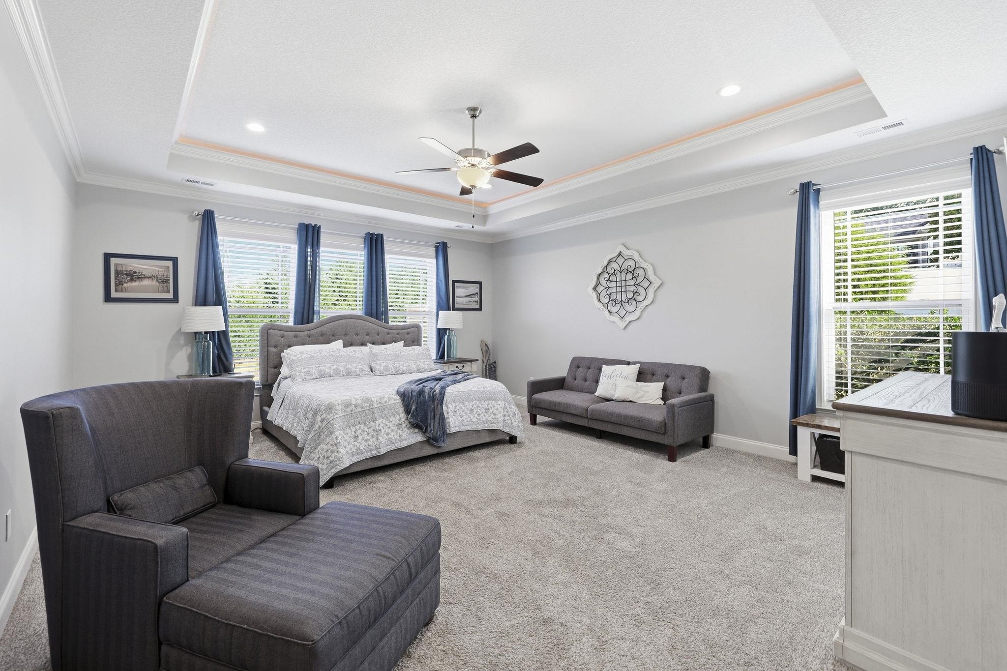 Furnished bedroom with blue curtains, a tray ceiling, and seating beside the bed.