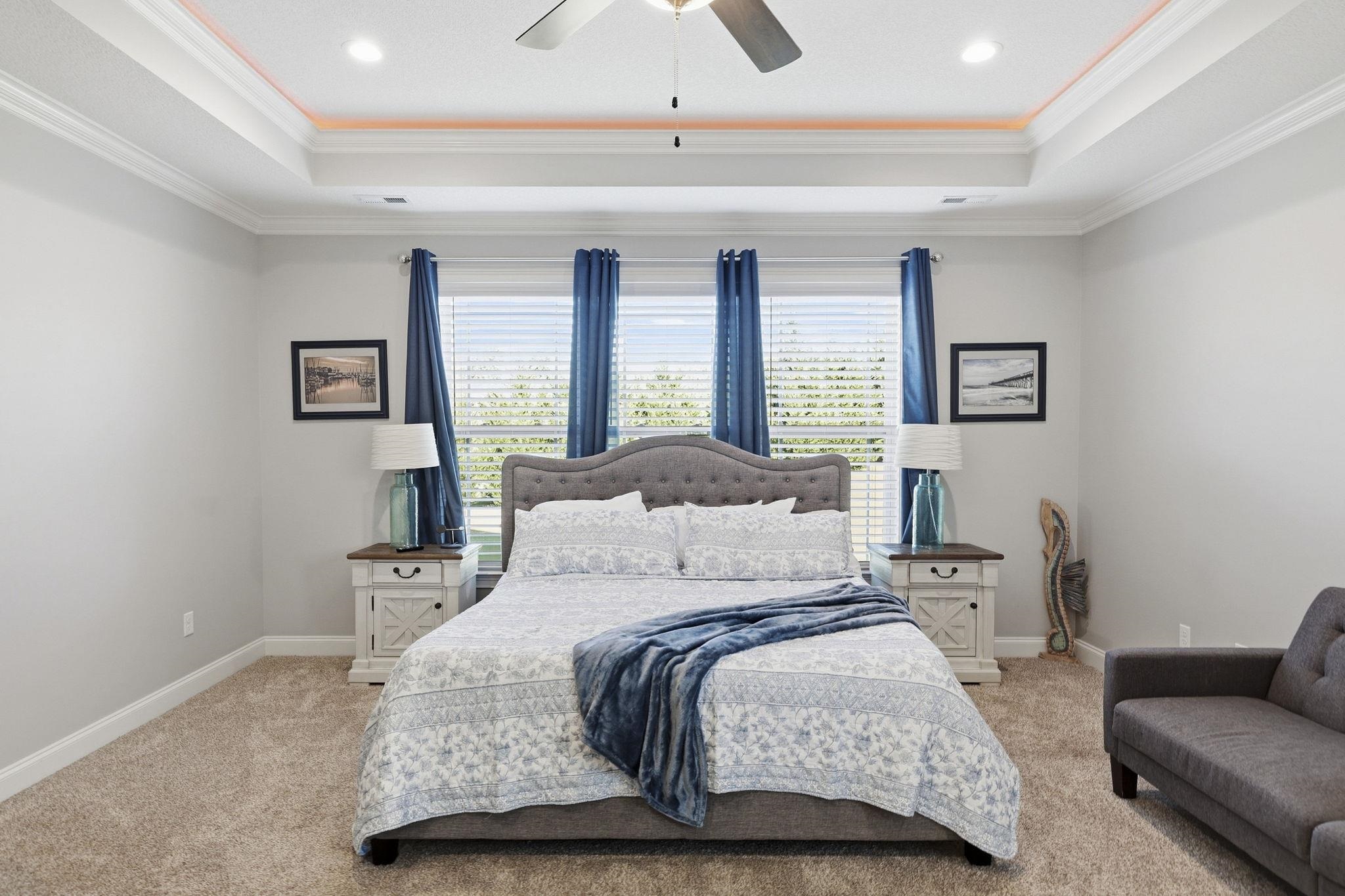 Bedroom with a centered upholstered bed, blue curtains, and a tray ceiling with orange accent lighting.