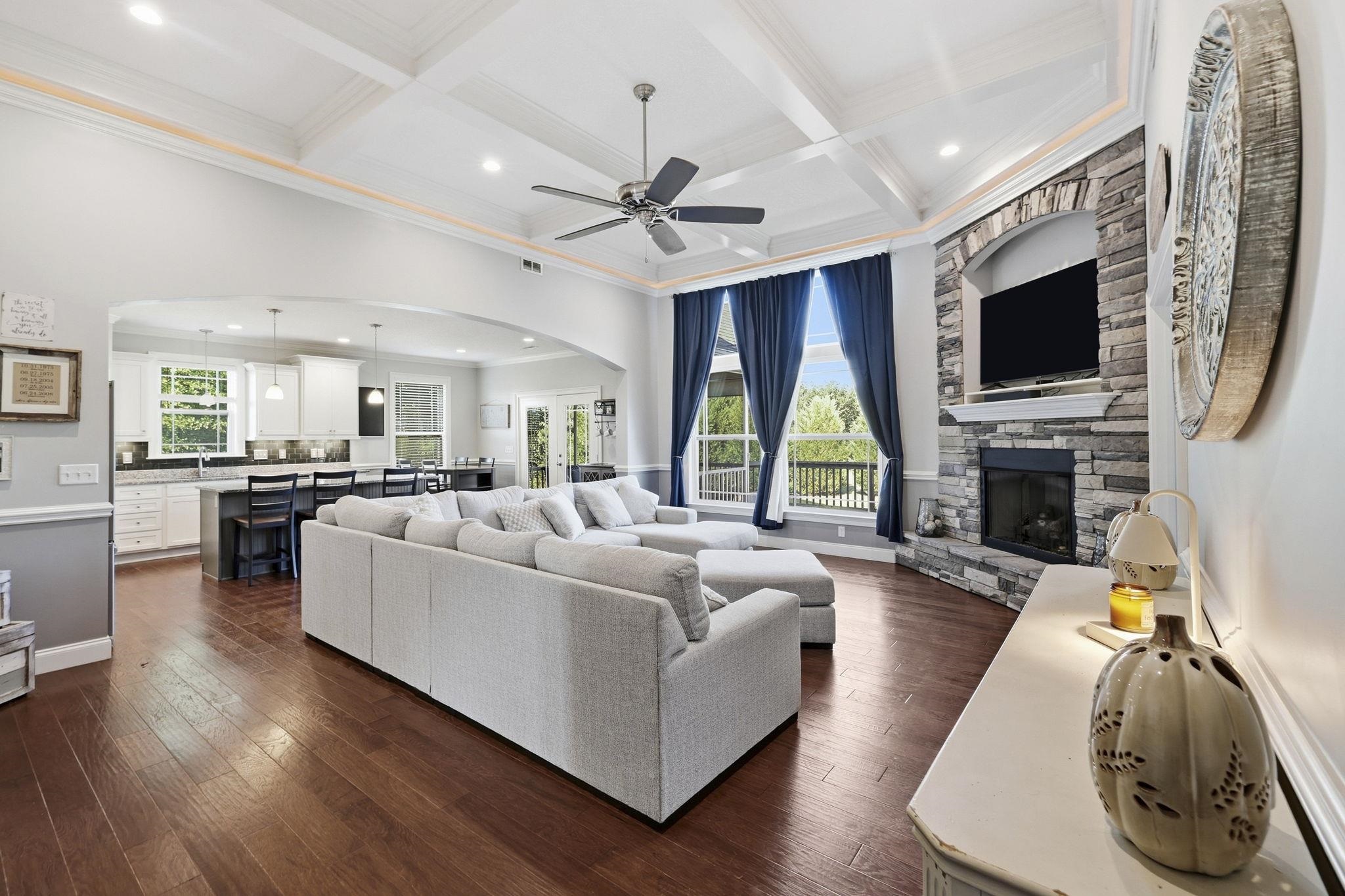 Living room with a gray sectional, tall blue-curtained windows, and a stone fireplace beside the open kitchen.
