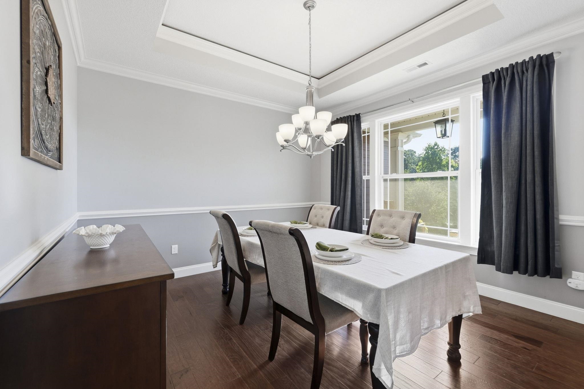 Dining room with a set table, tray ceiling, chandelier, and large curtained windows.