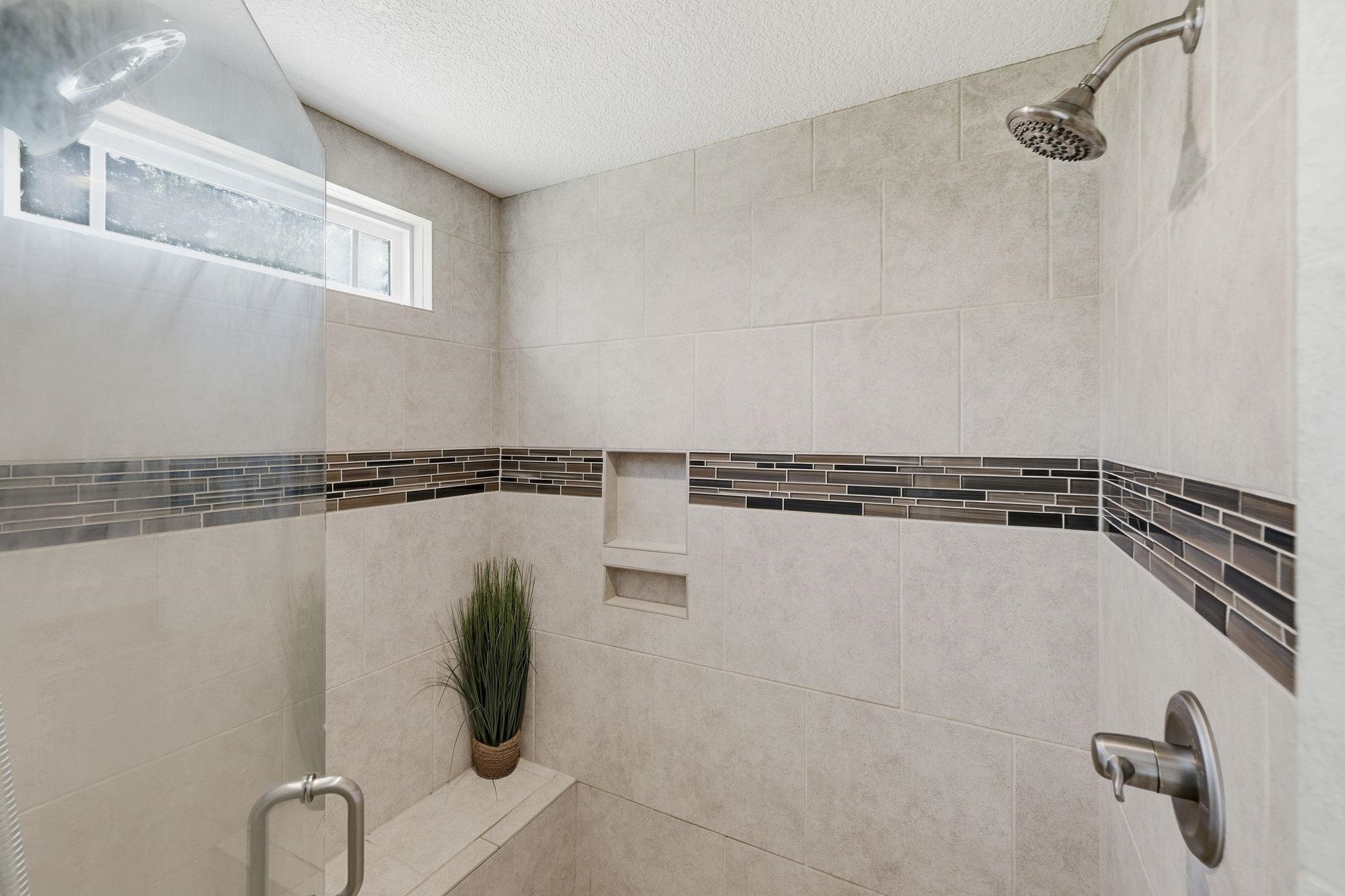 Walk-in shower with beige tile, dark mosaic accent stripe, built-in niches, and a tiled bench with a potted plant.