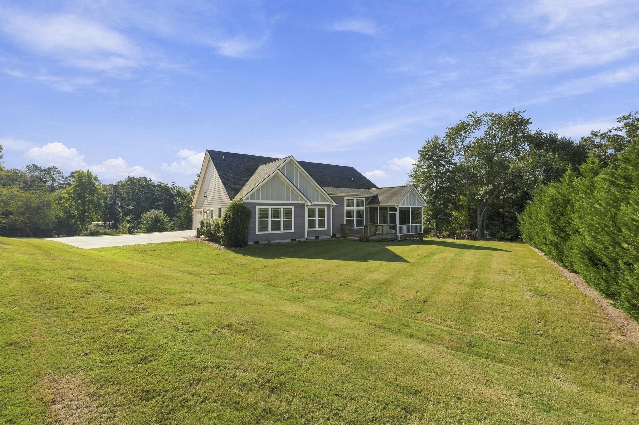 Gray gabled house with a covered deck beyond a broad lawn bordered by trees and hedges.