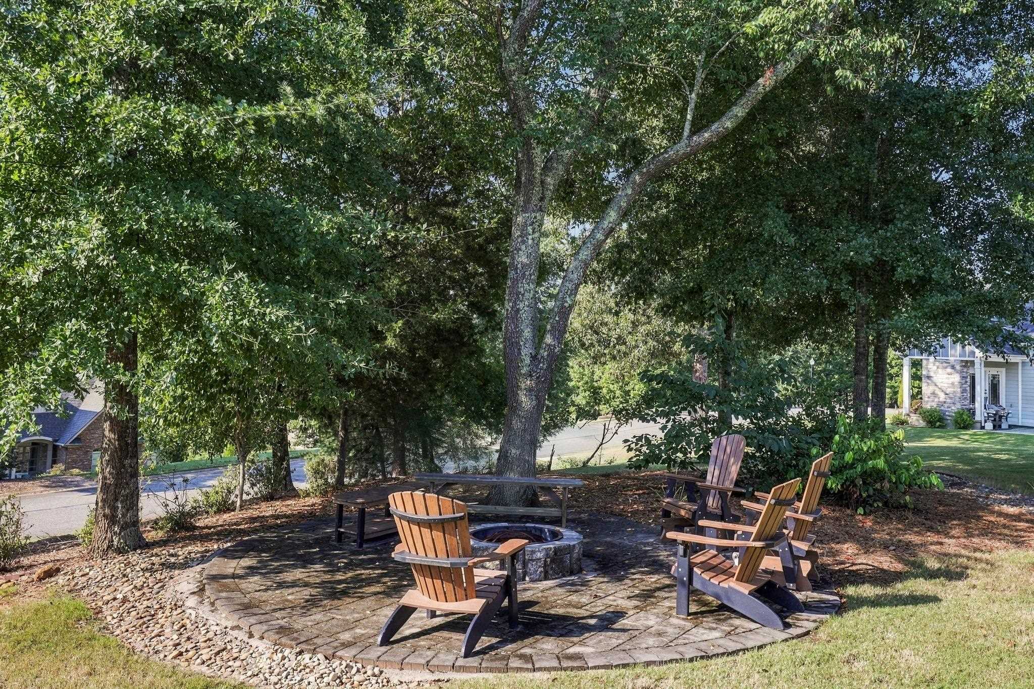 Shaded patio with a stone fire pit and Adirondack chairs beneath mature trees.