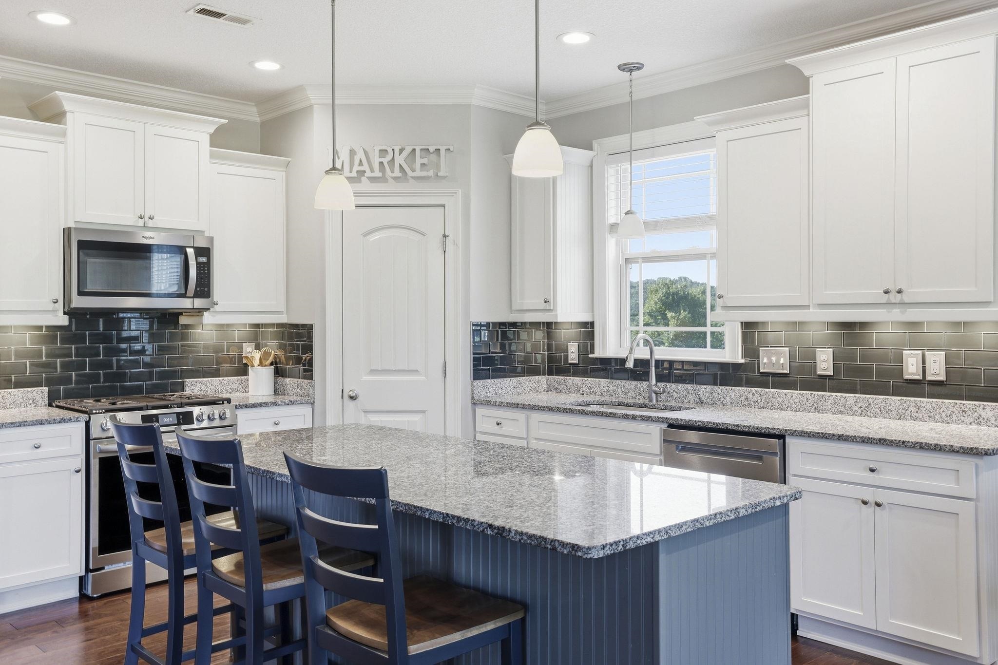 Kitchen with a blue island and bar stools, white cabinets, dark tile backsplash, and a window over the sink.