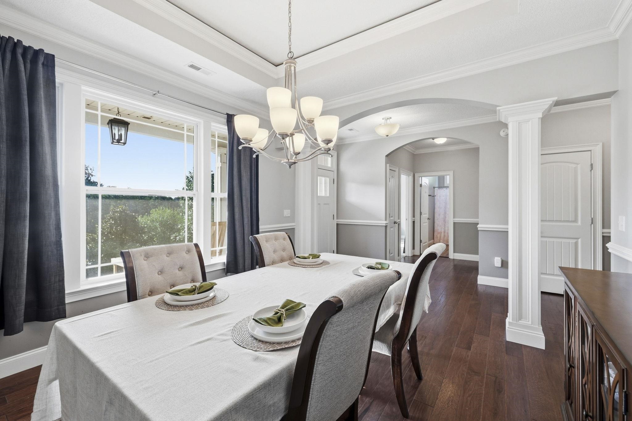 Formal dining room with a set table beneath a chandelier and a large curtained window.