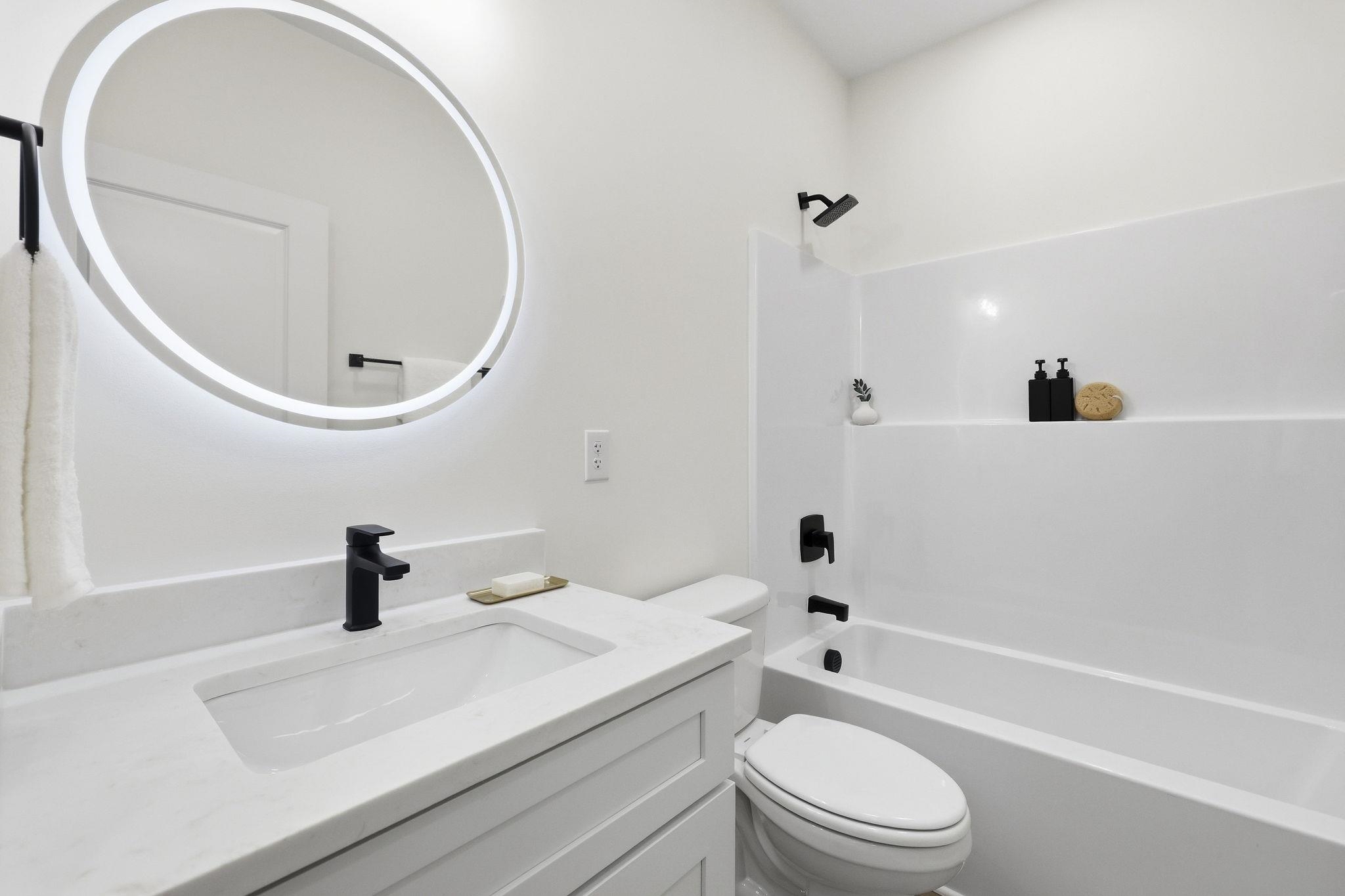 Bright white bathroom with a round illuminated mirror, black fixtures, vanity, toilet, and tub-shower.