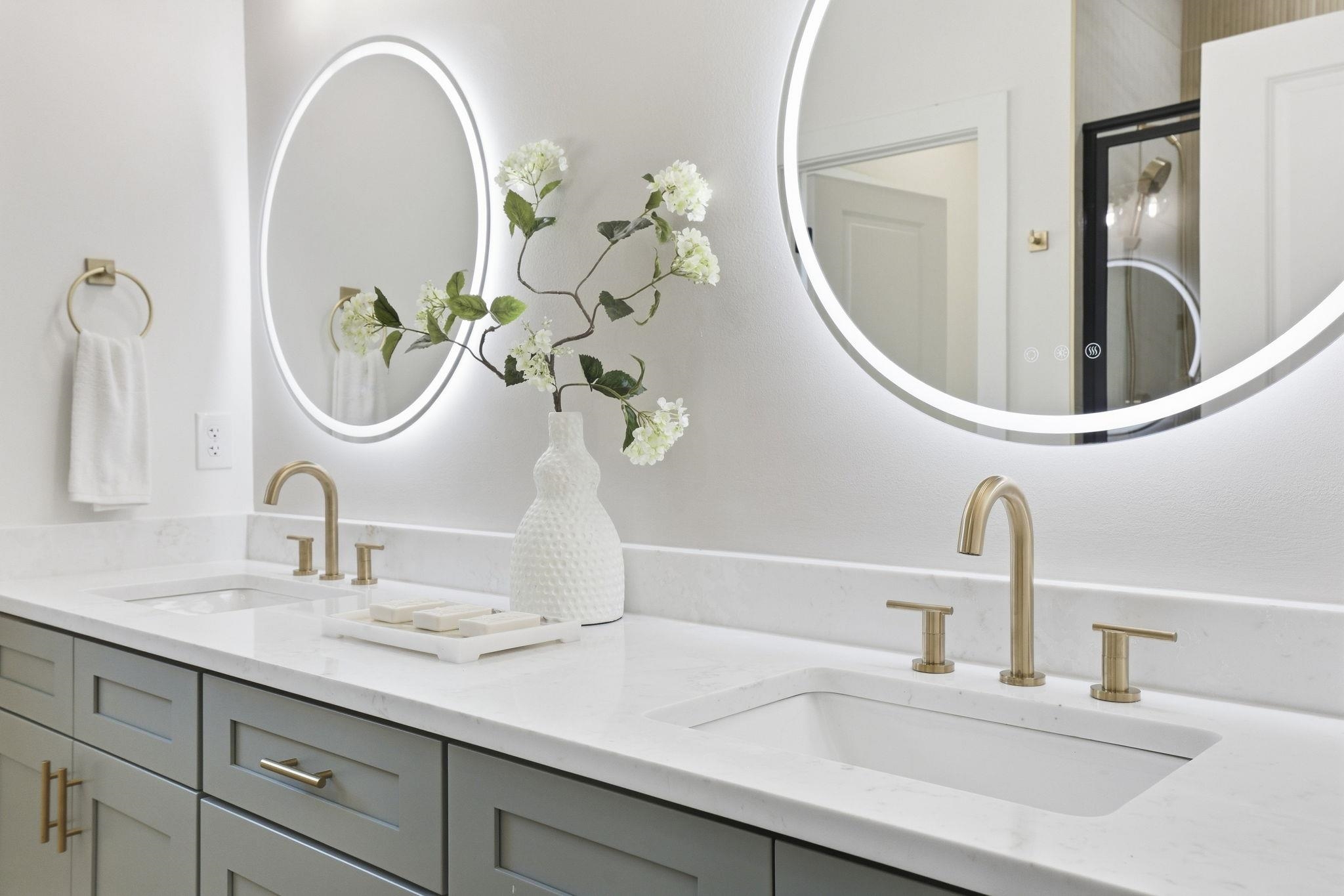 Bathroom vanity with double sinks, gold faucets, gray cabinets, and illuminated round mirrors.