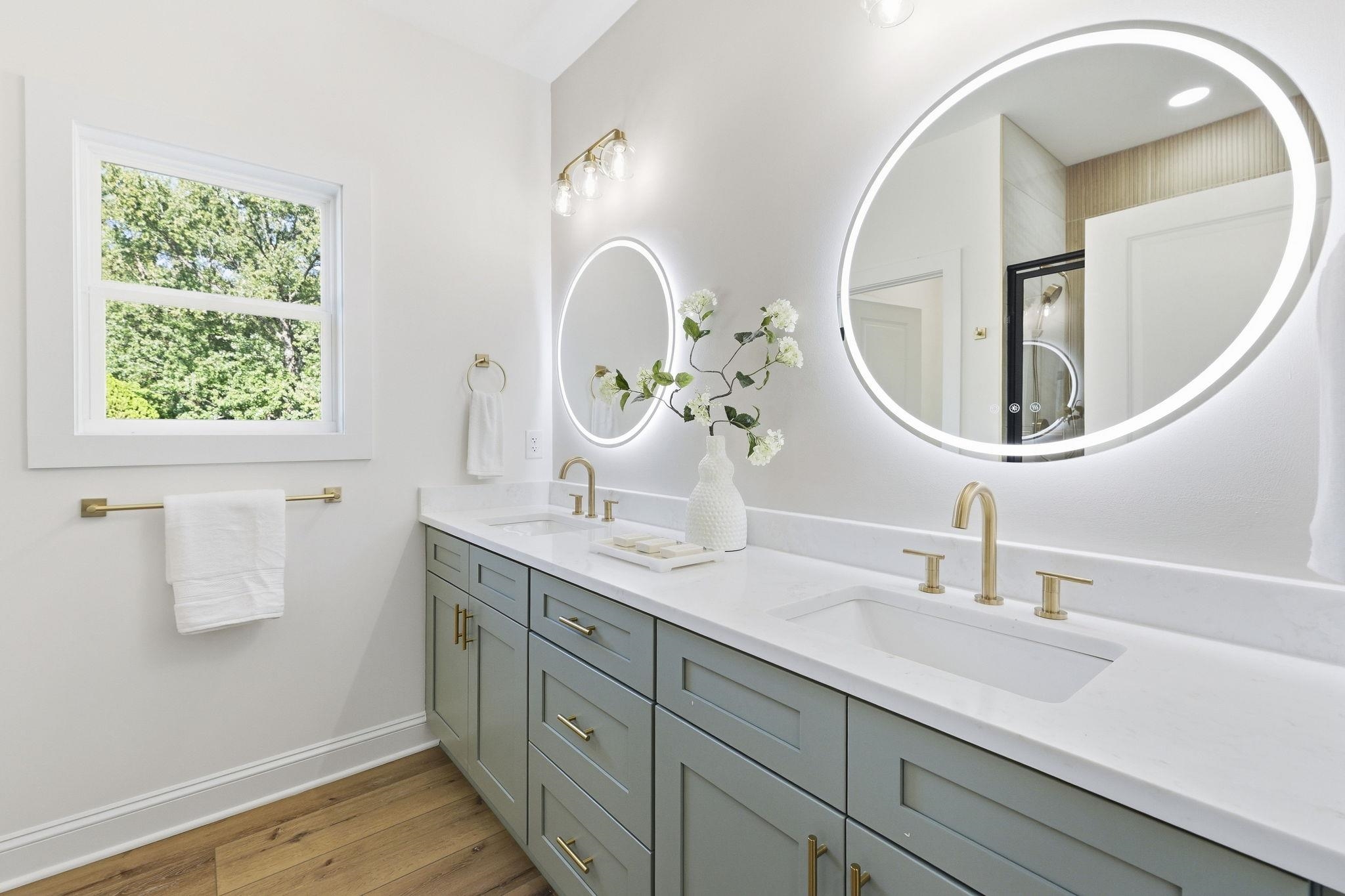 Bright bathroom with a sage-green double vanity, brass fixtures, and large illuminated round mirrors.