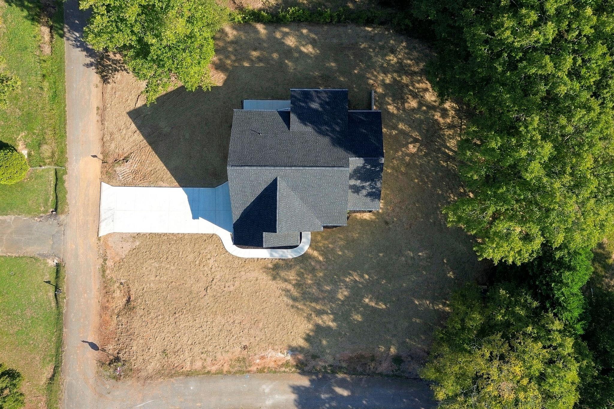 Aerial view of a dark-roofed house, concrete driveway, and partly cleared yard surrounded by trees.