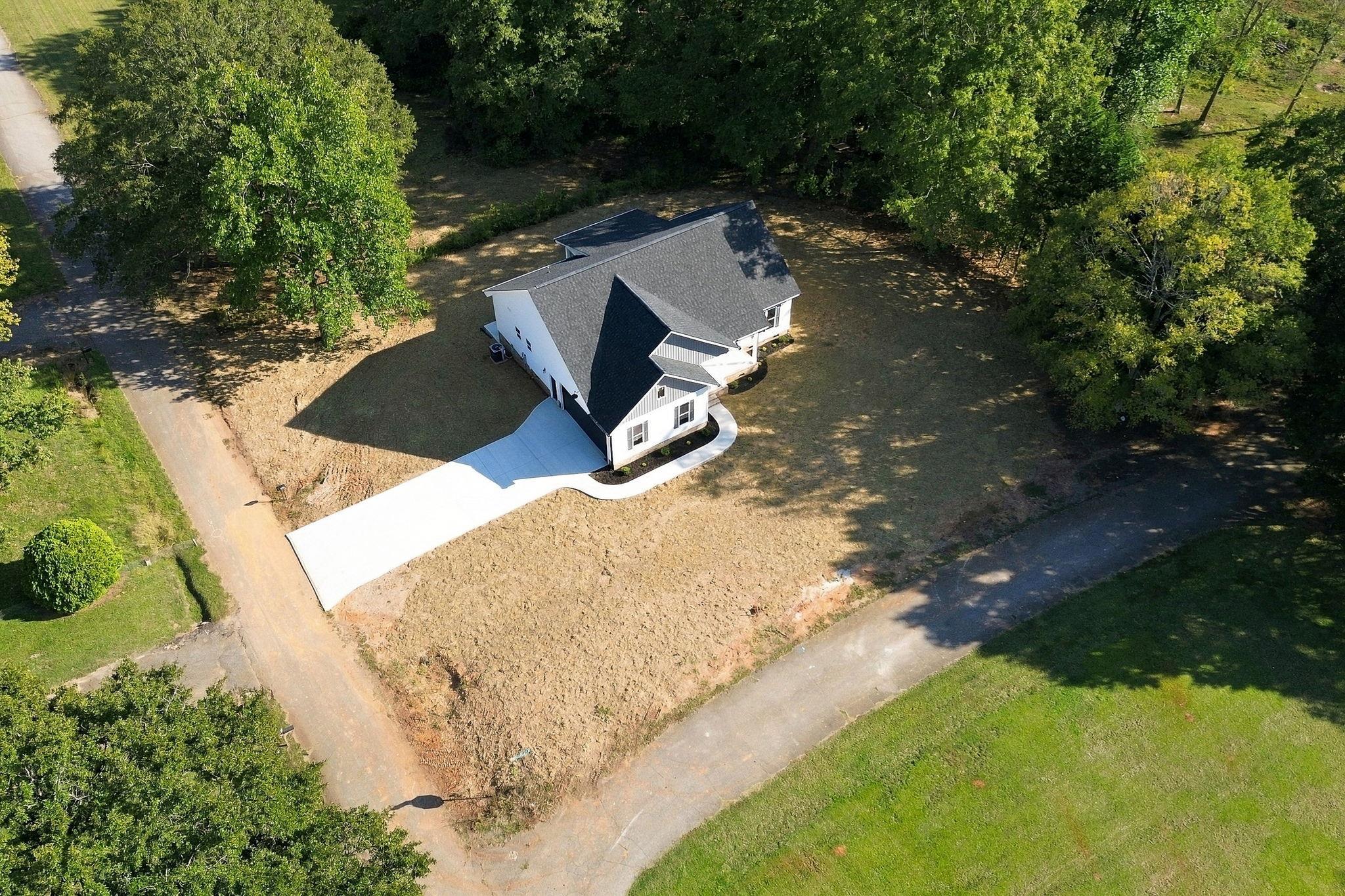 Aerial view of a white house with a dark gabled roof, wide driveway, and wooded lot.