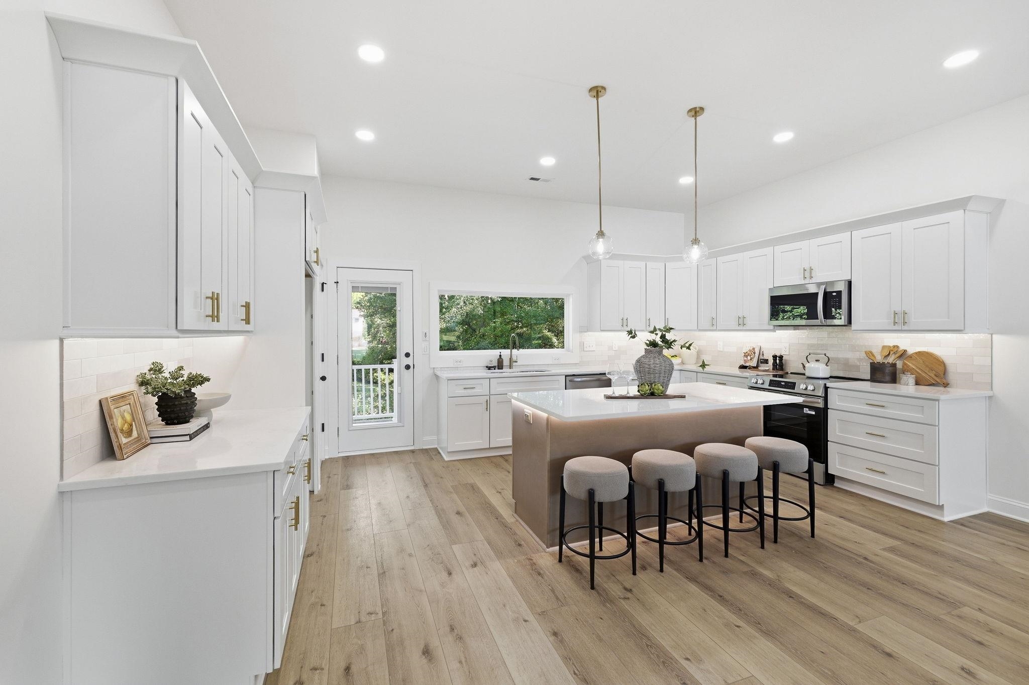 Modern white kitchen with a large island, four stools, brass hardware, and a window over the sink.