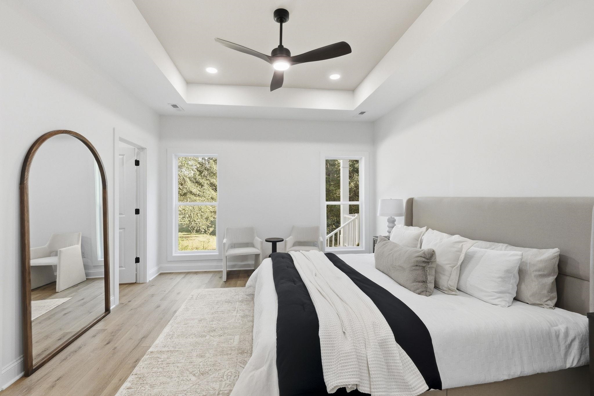Staged bedroom with a tray ceiling, dark ceiling fan, arched mirror, and windows overlooking greenery.