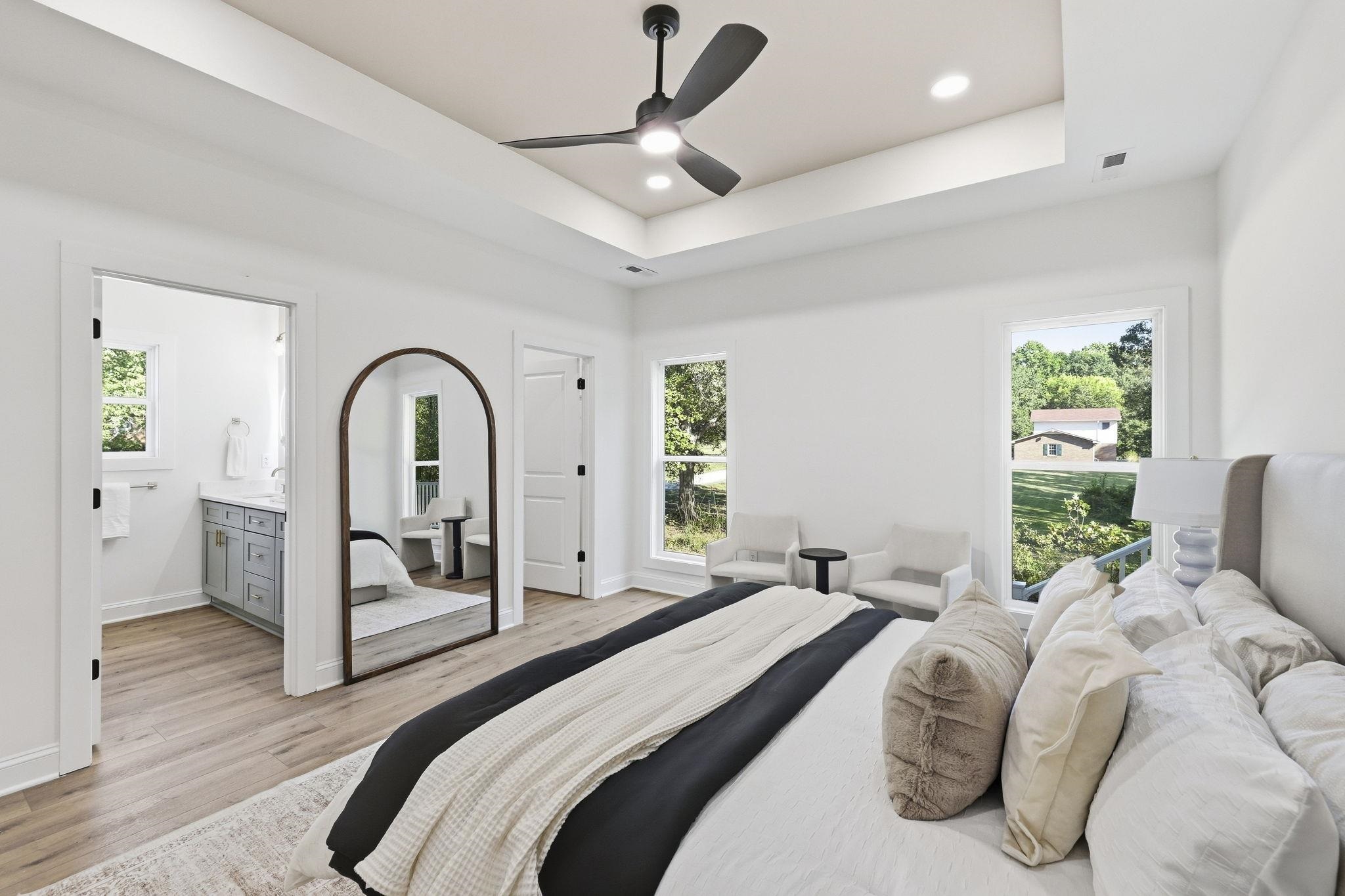 Staged bedroom with tray ceiling, black fan, arched mirror, and doorway to an adjoining bathroom.