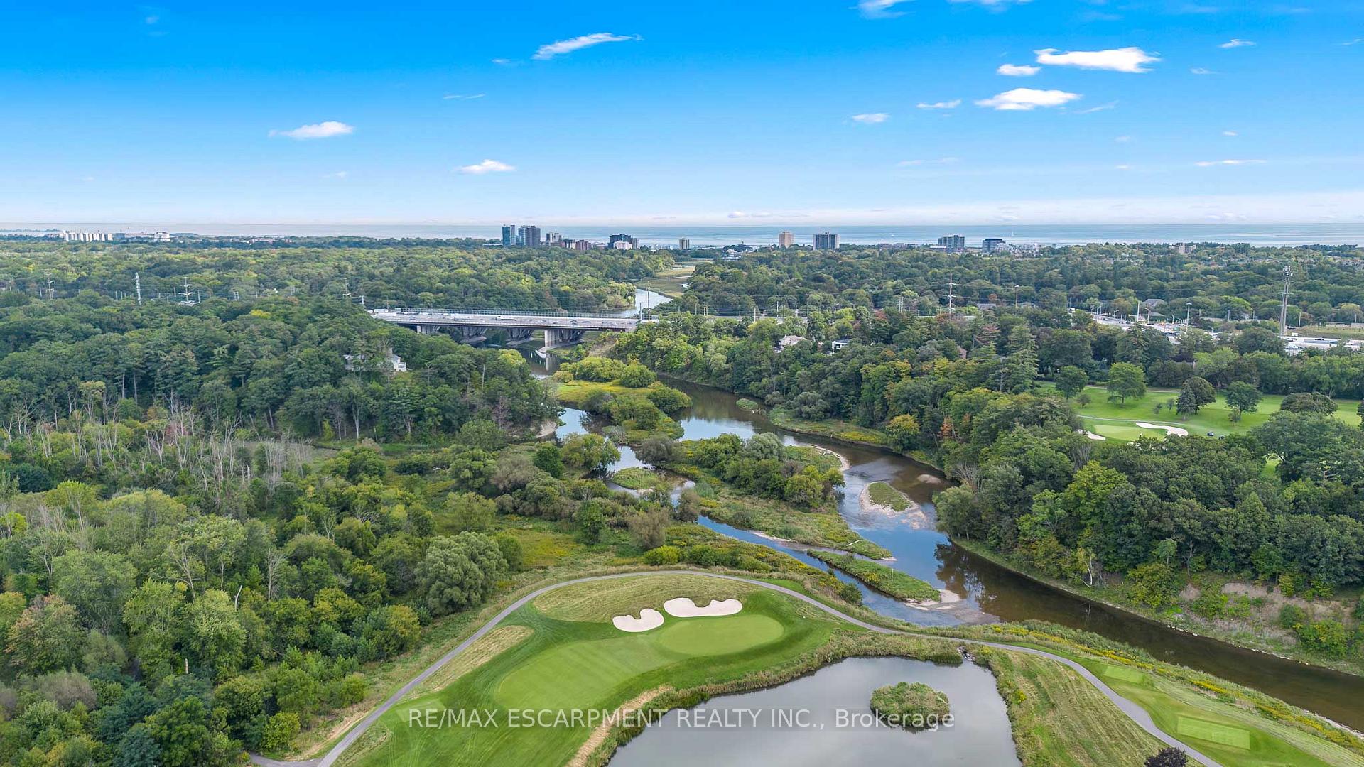 Aerial view of a wooded river and bridge beside a golf course, with distant buildings on the horizon.