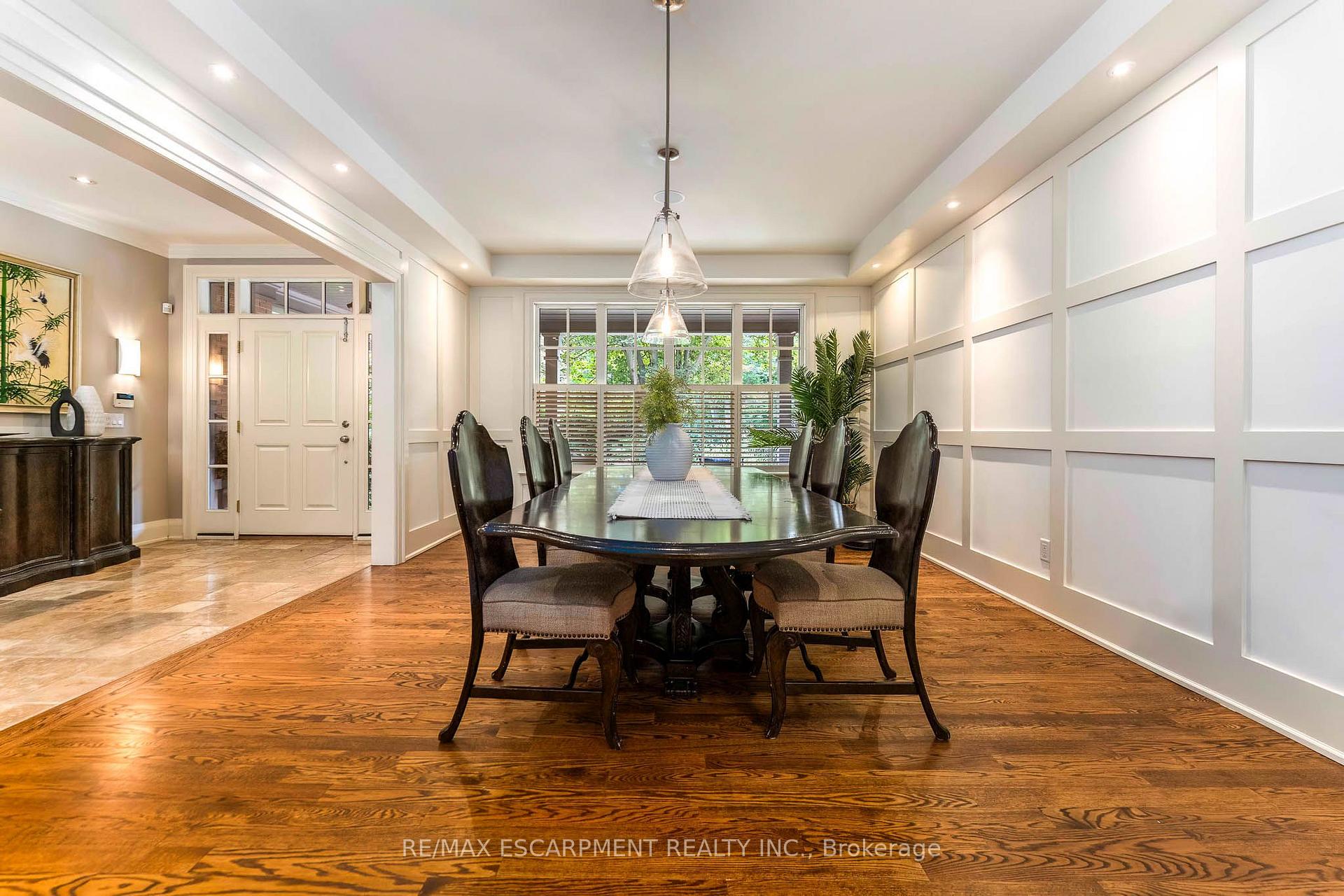 Dining room with a dark wood table, paneled walls, hardwood floors, and windows overlooking greenery.