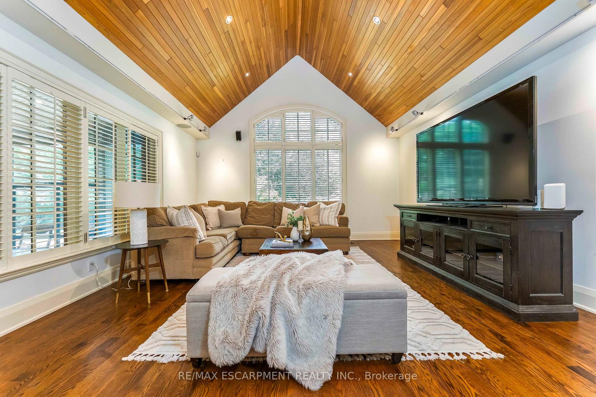 Living room with a vaulted wood ceiling, brown sectional sofa, shuttered windows, and large television.