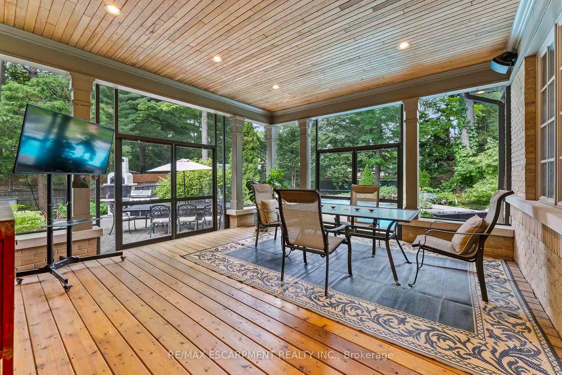 Enclosed sunroom with a wood ceiling, dining set, lounge chairs, and garden and pool views.