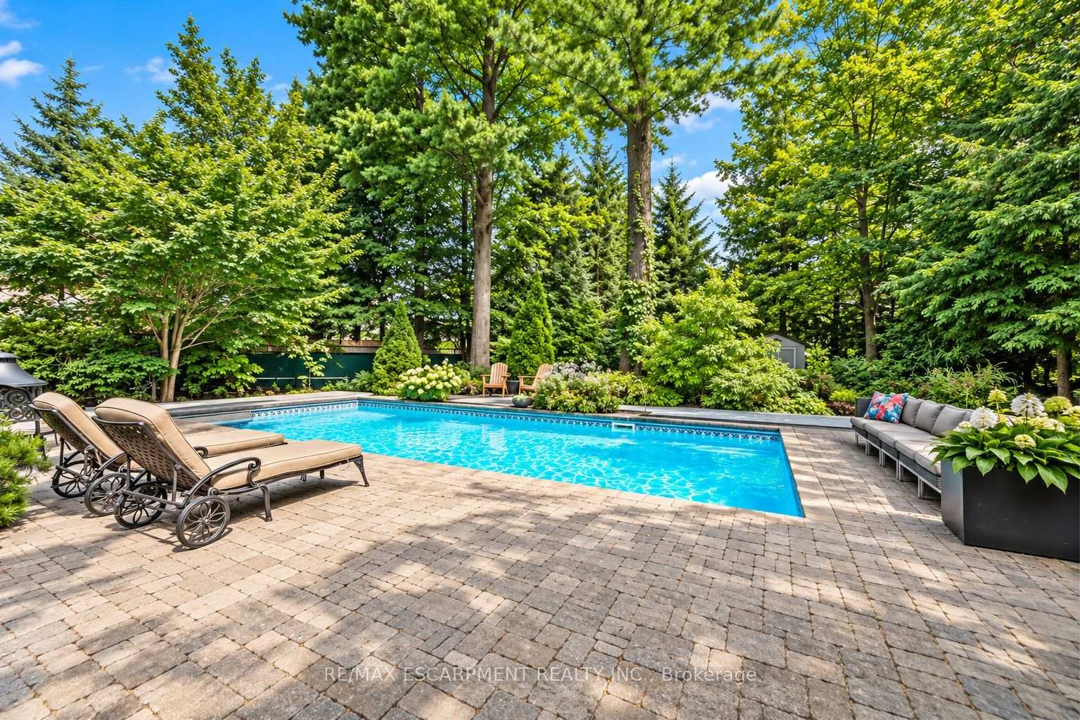 Backyard swimming pool with a stone patio, lounge chairs, and mature trees.