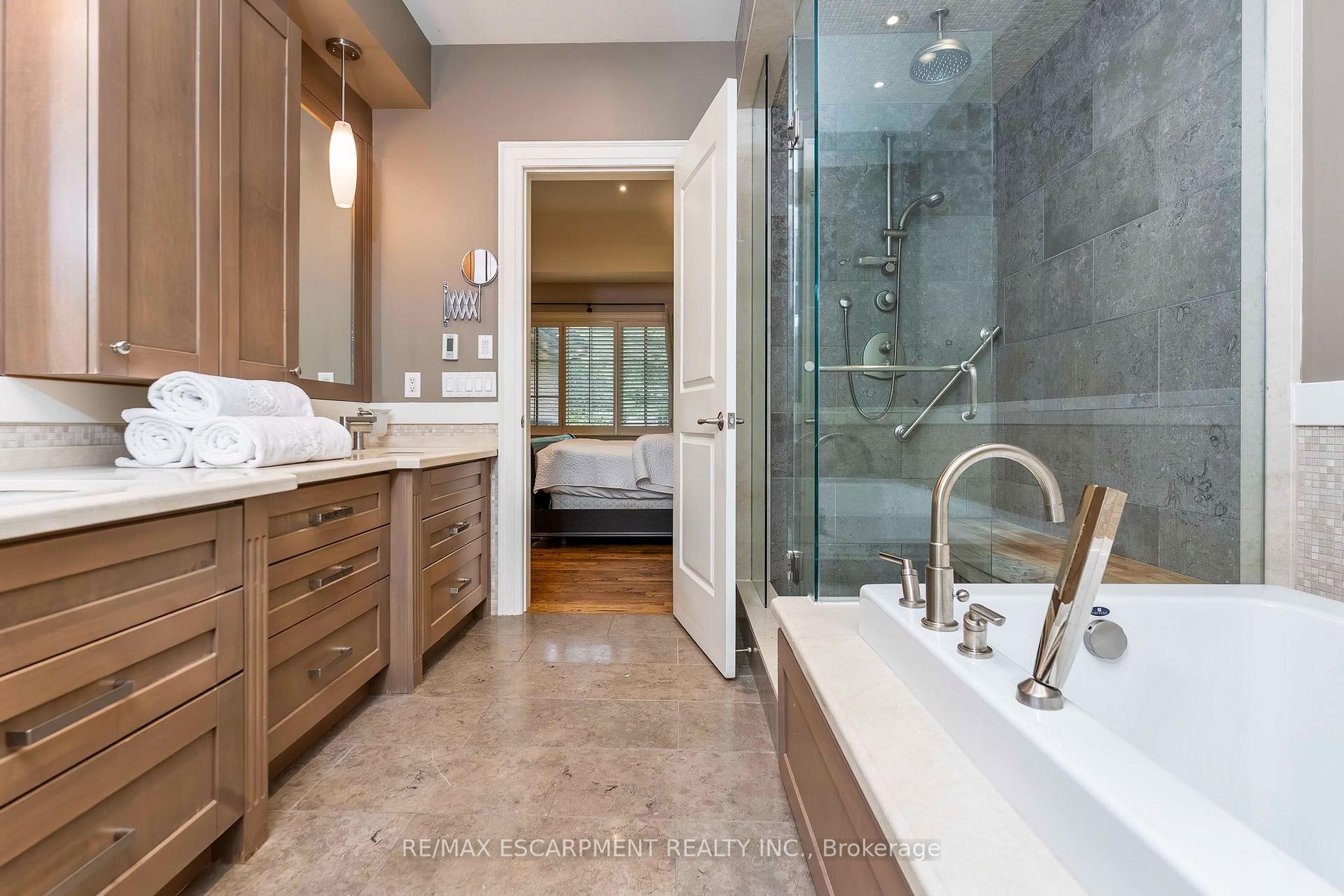 Primary bathroom with a gray-tiled glass shower, soaking tub, and wood vanity; bedroom visible through the doorway.