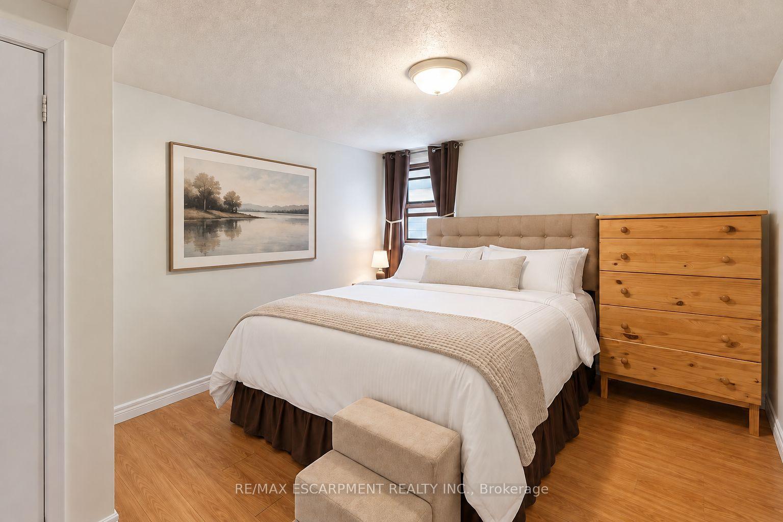 Staged bedroom with a large white bed, tufted headboard, wooden dresser, and hardwood floor.