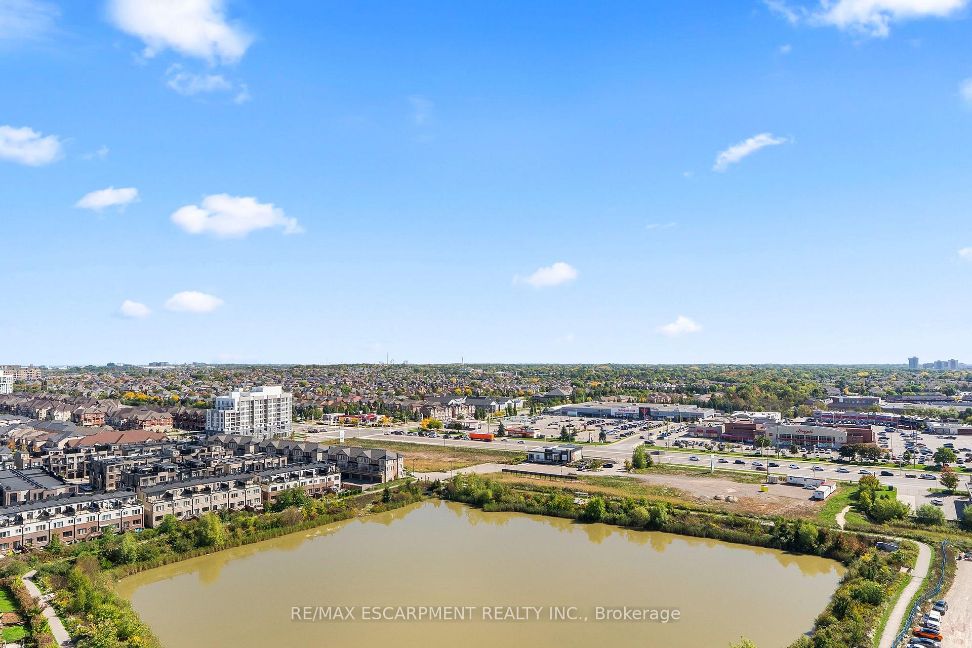 Aerial view of a pond, townhouses, and surrounding Oakville neighborhoods beneath a broad blue sky.