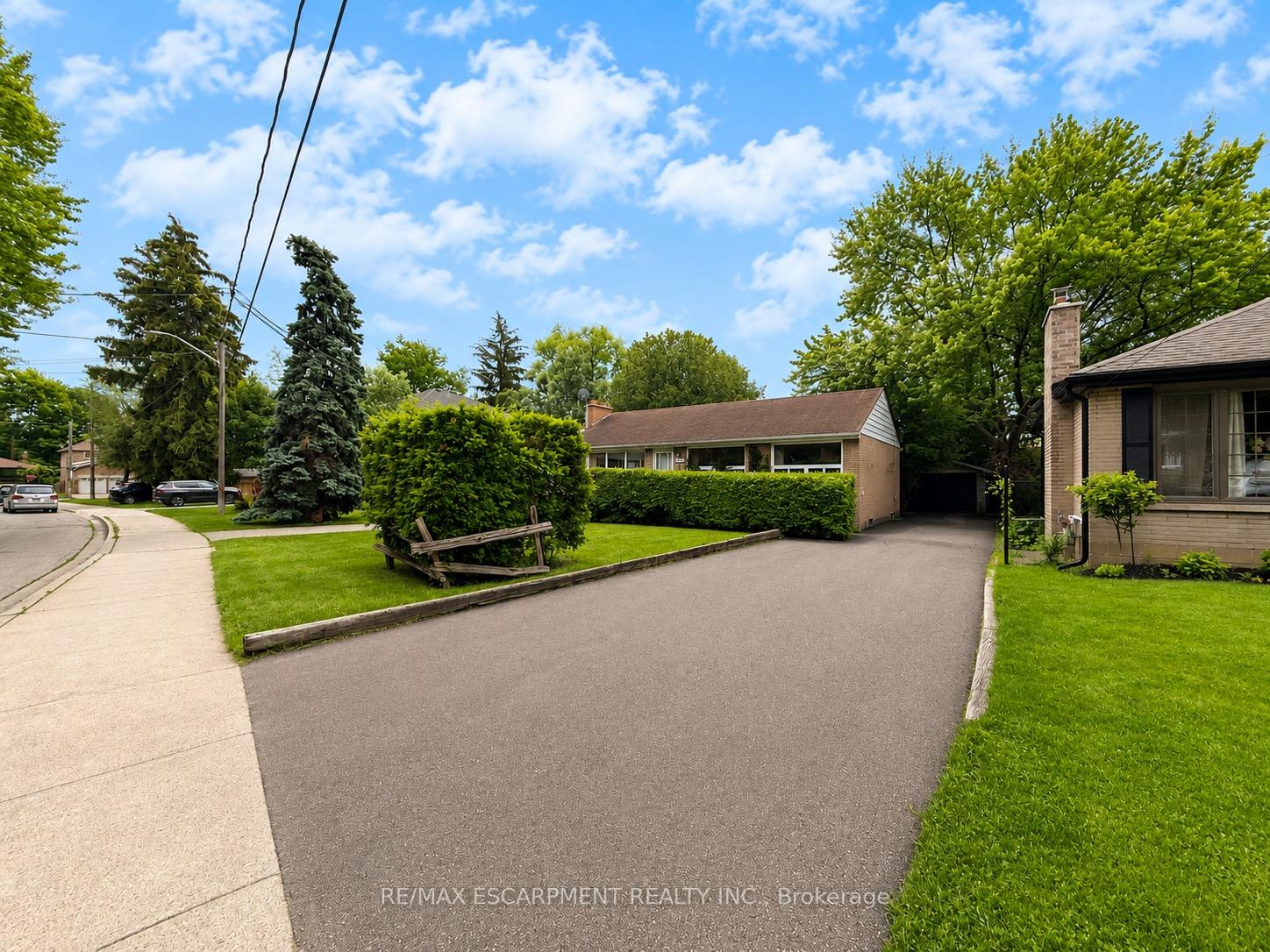 Brick home with a broad driveway, trimmed hedges, mature trees, and a wooden garden bench.