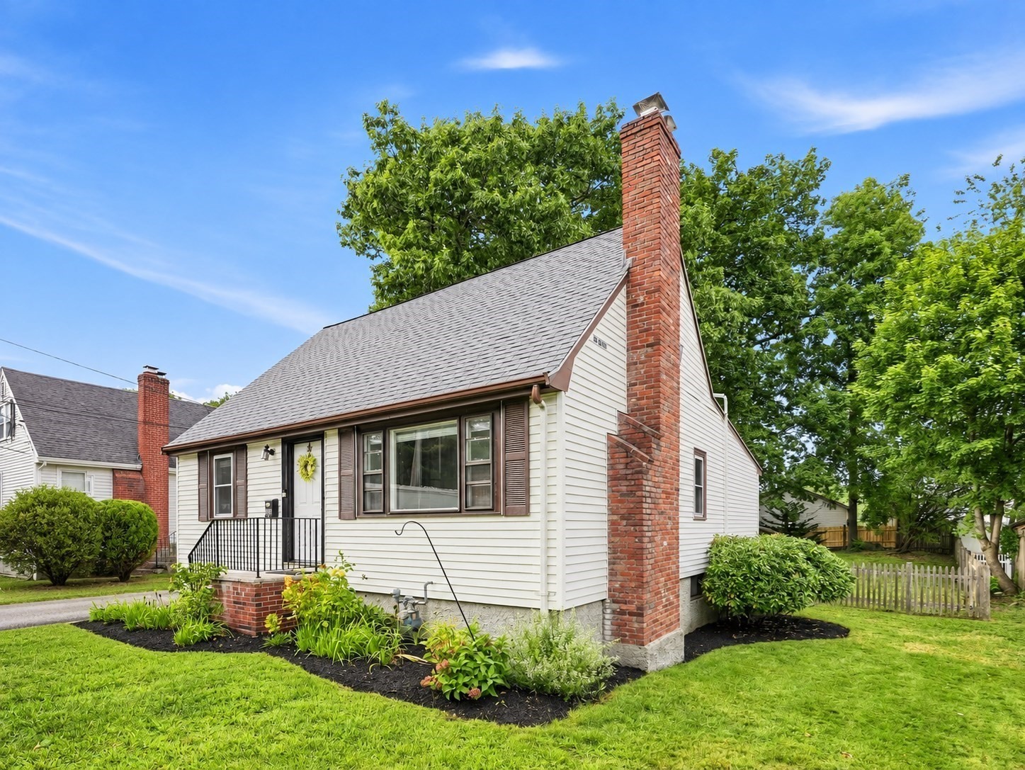 White-sided house with a steep gray roof, tall brick chimney, and landscaped front lawn.