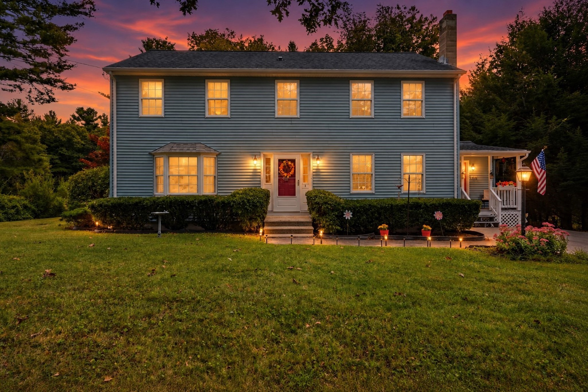 Gray two-story house with warmly lit windows, a bay window, and a covered porch at sunset.