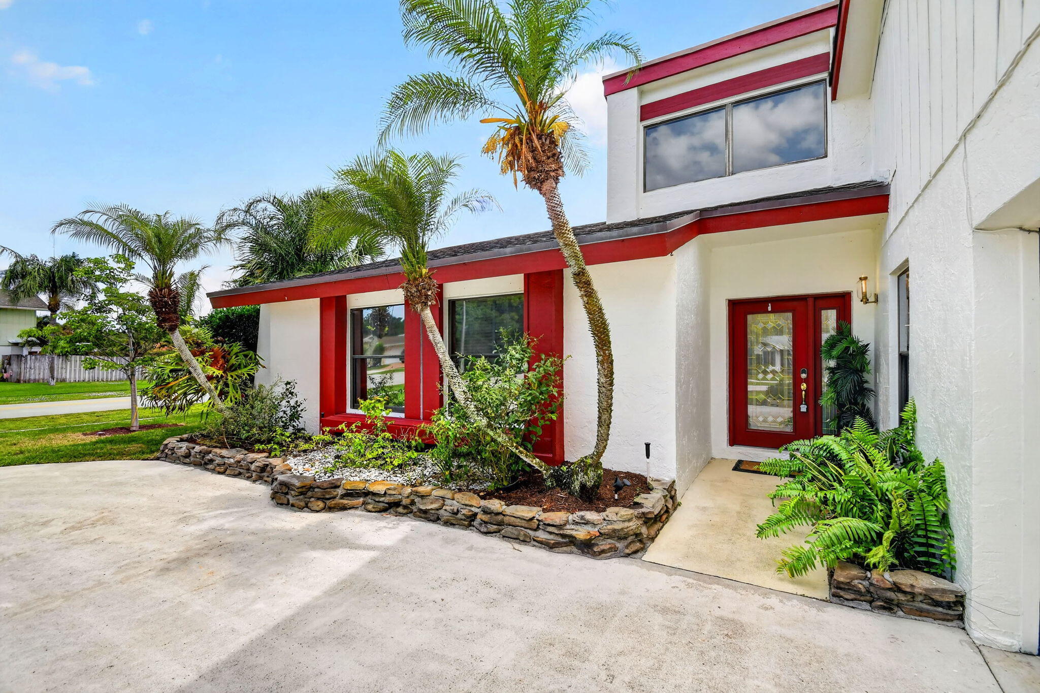White two-story home with red trim and entry, framed by palms and landscaped stone beds.