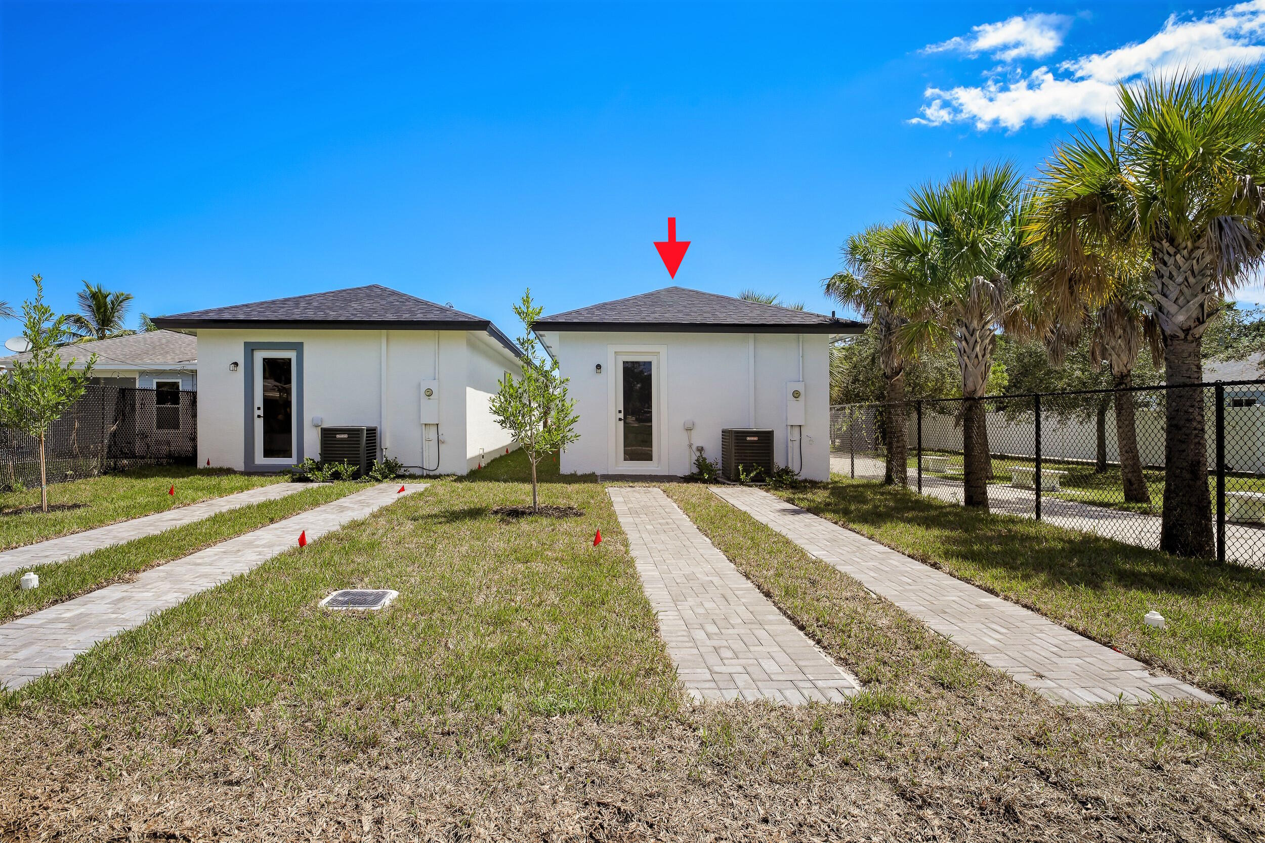 White single-story home with a dark hip roof, marked by a red arrow beside a paved walkway and palm trees.