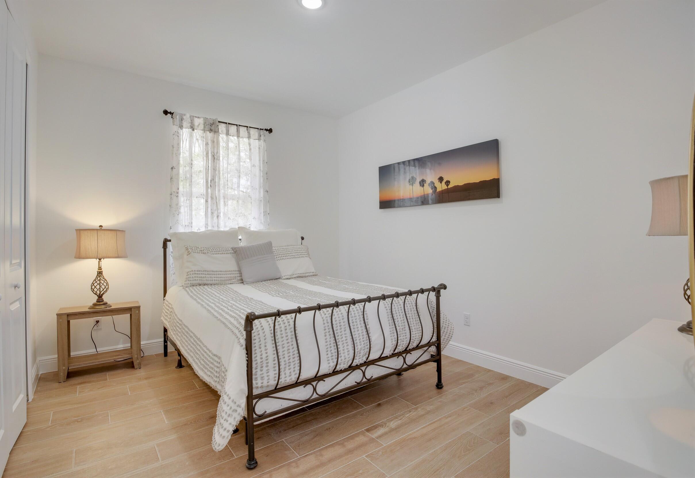 Bedroom with a dark metal bed, white textured bedding, sheer curtains, and wood-look tile flooring.