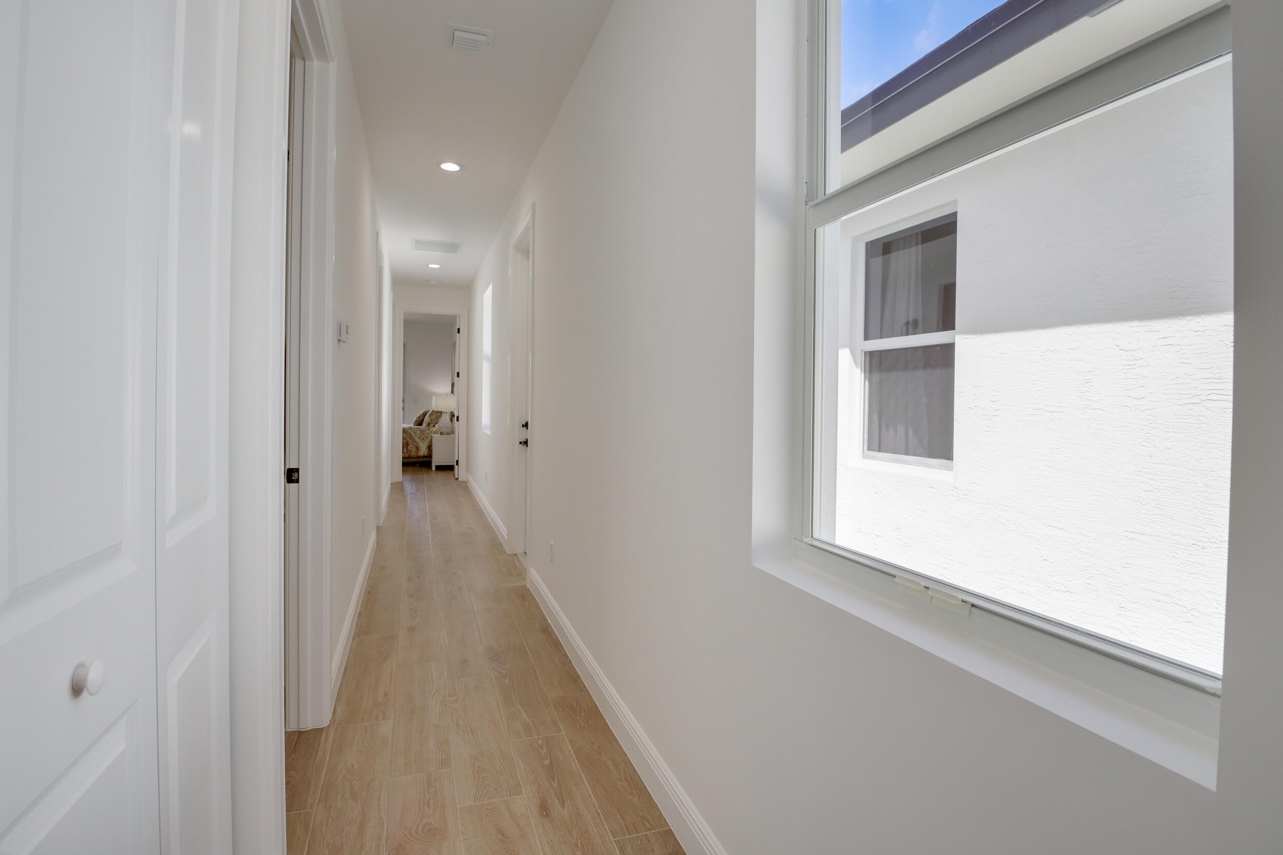 Bright hallway with wood-look tile flooring and a large window, leading to a furnished bedroom.