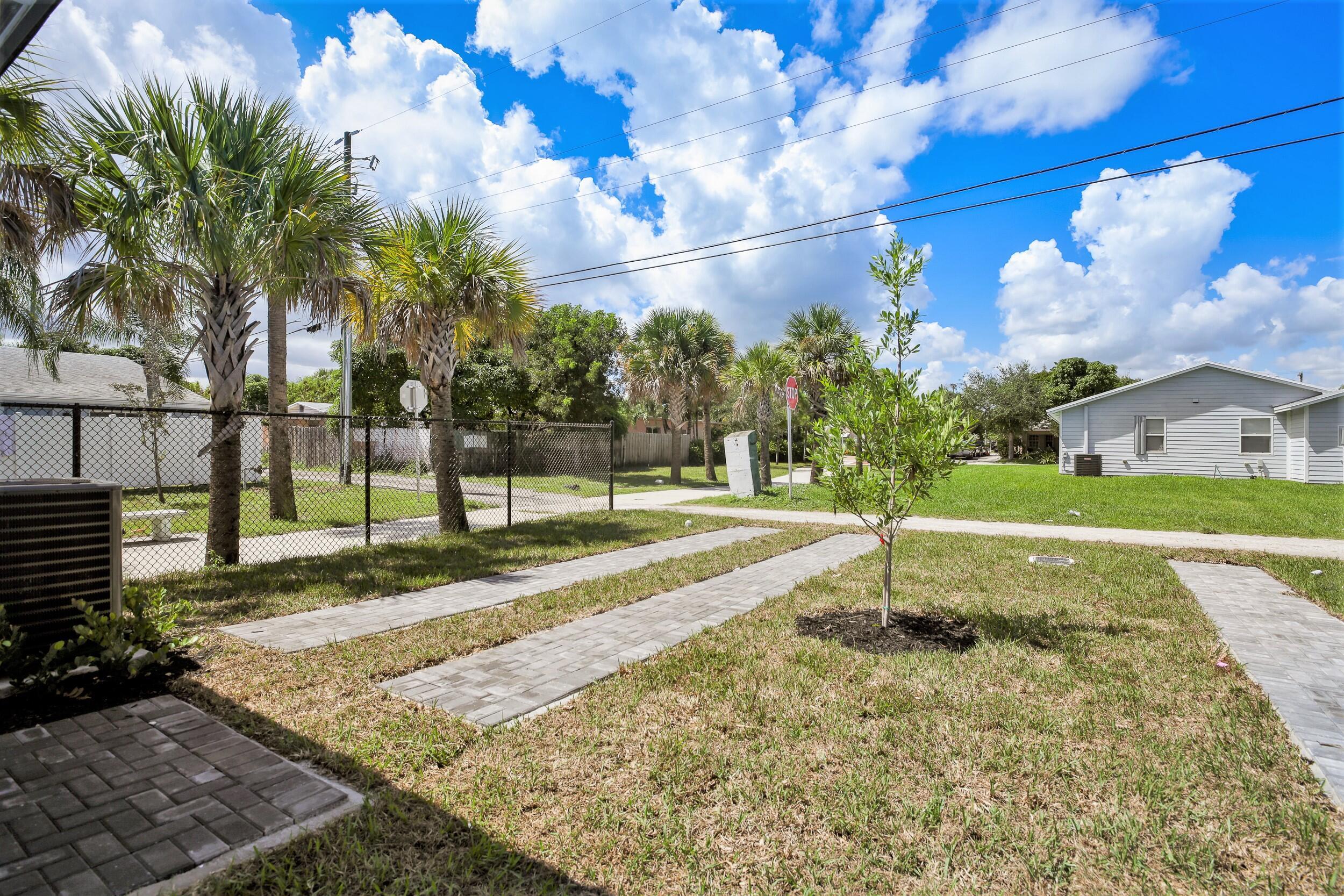 Residential yard with paver walkways, a young tree, palm trees, and a light gray house.