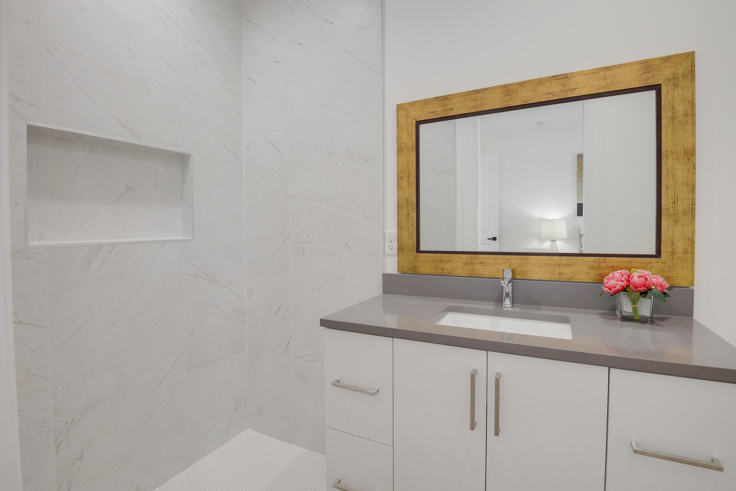 Modern bathroom vanity with a gold-framed mirror, gray countertop, and marble-look tiled shower niche.