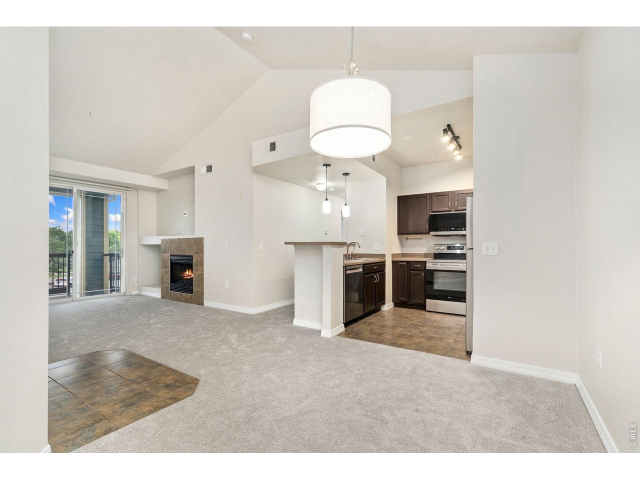 Vacant living area with vaulted ceilings, tiled fireplace, balcony doors, and an open kitchen.