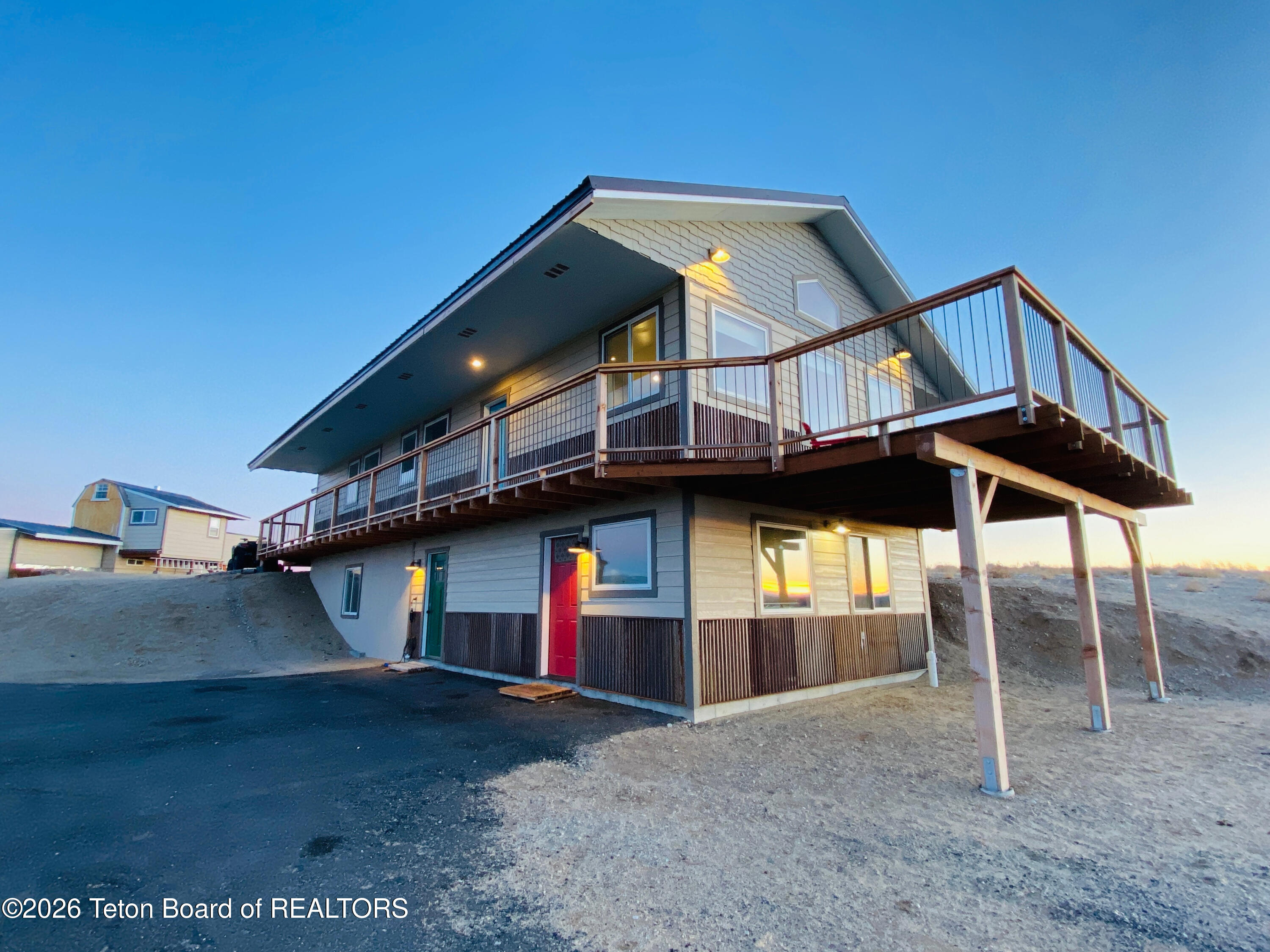 Two-story house with a large elevated wraparound deck on a sandy lot at dusk.